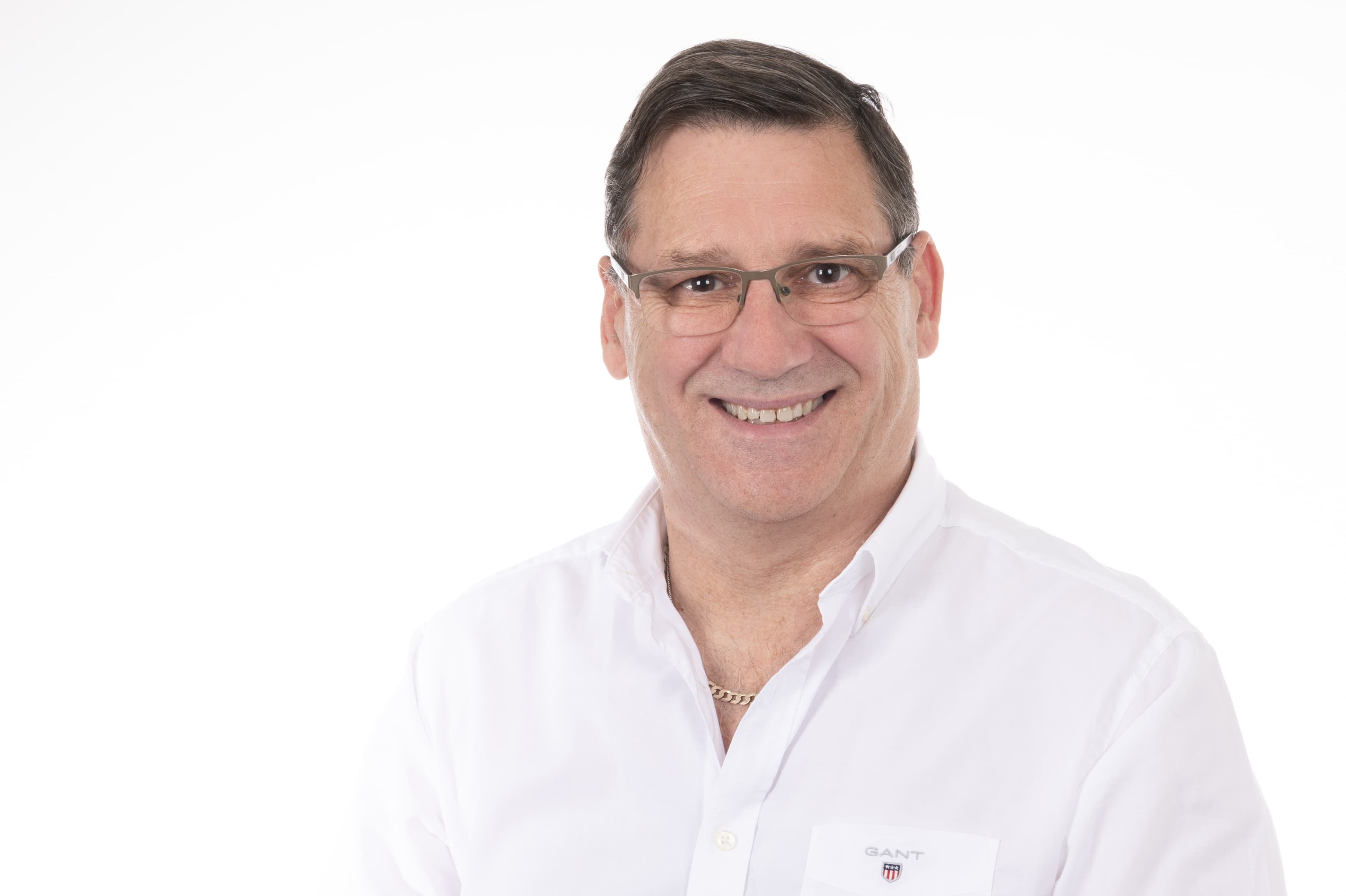 Smiling person with short dark hair and glasses, wearing a white button-down shirt with a gold chain, and a small GANT badge on the pocket.