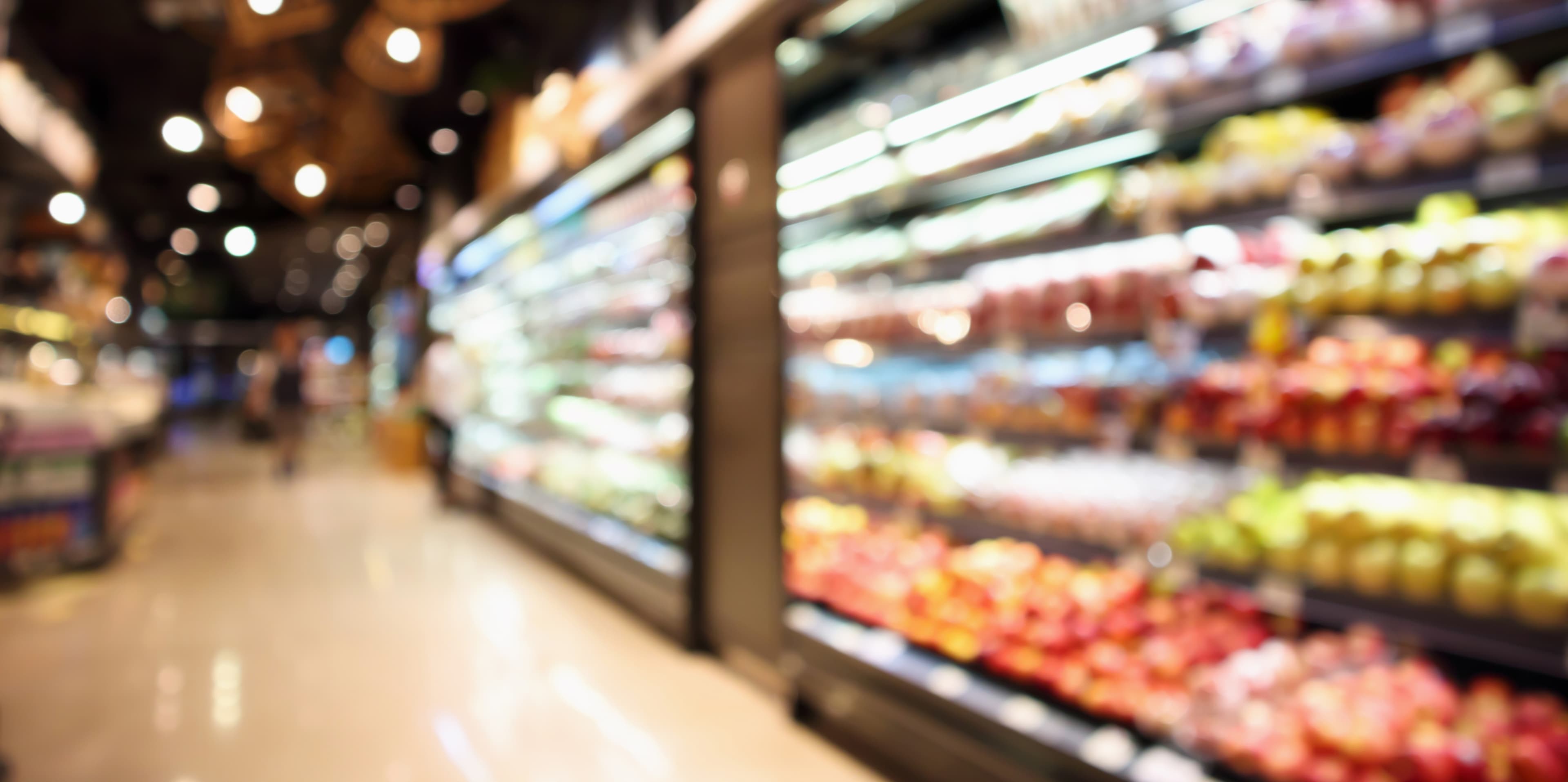 Blurred supermarket interior with bright overhead lights, a long refrigerated display on the right, and colorful produce shelves.