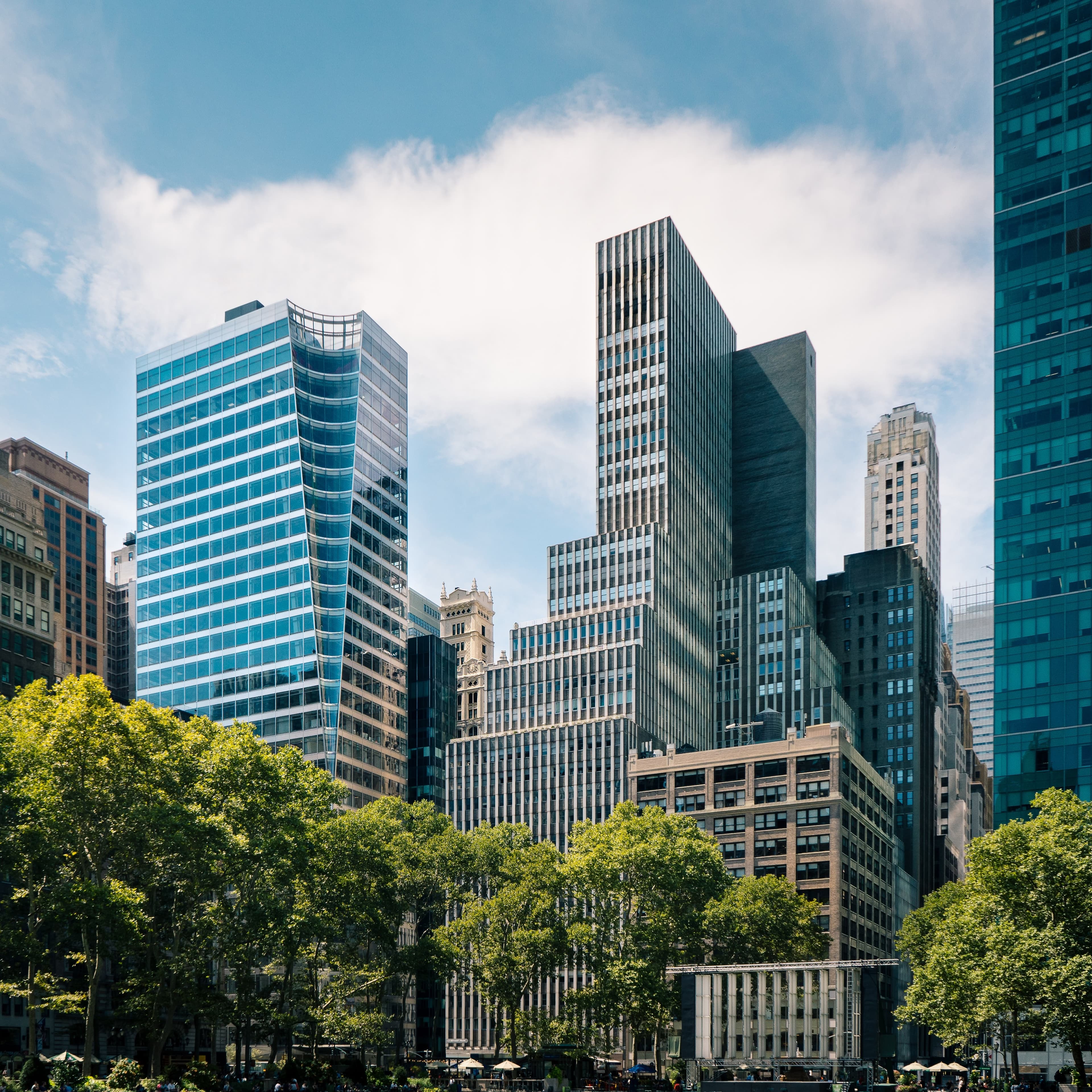 Paysage urbain avec des gratte-ciel modernes en verre, arbres au premier plan, ciel bleu et quelques nuages.