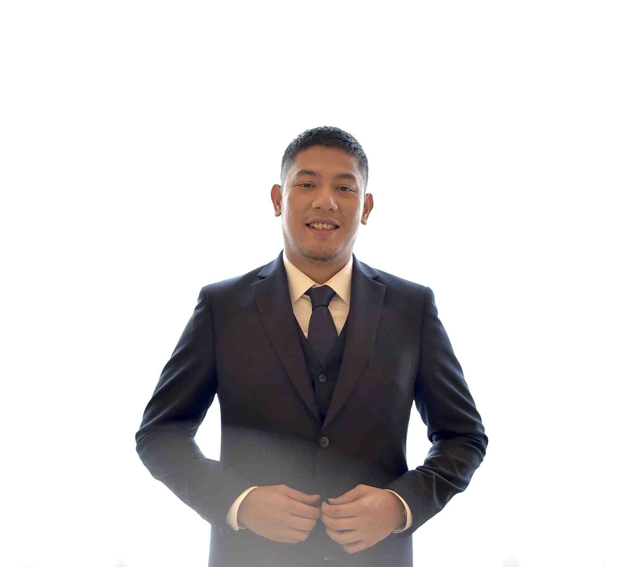 Person in a dark suit with a vest and navy tie, smiling with hands clasped at the waist against a white background.