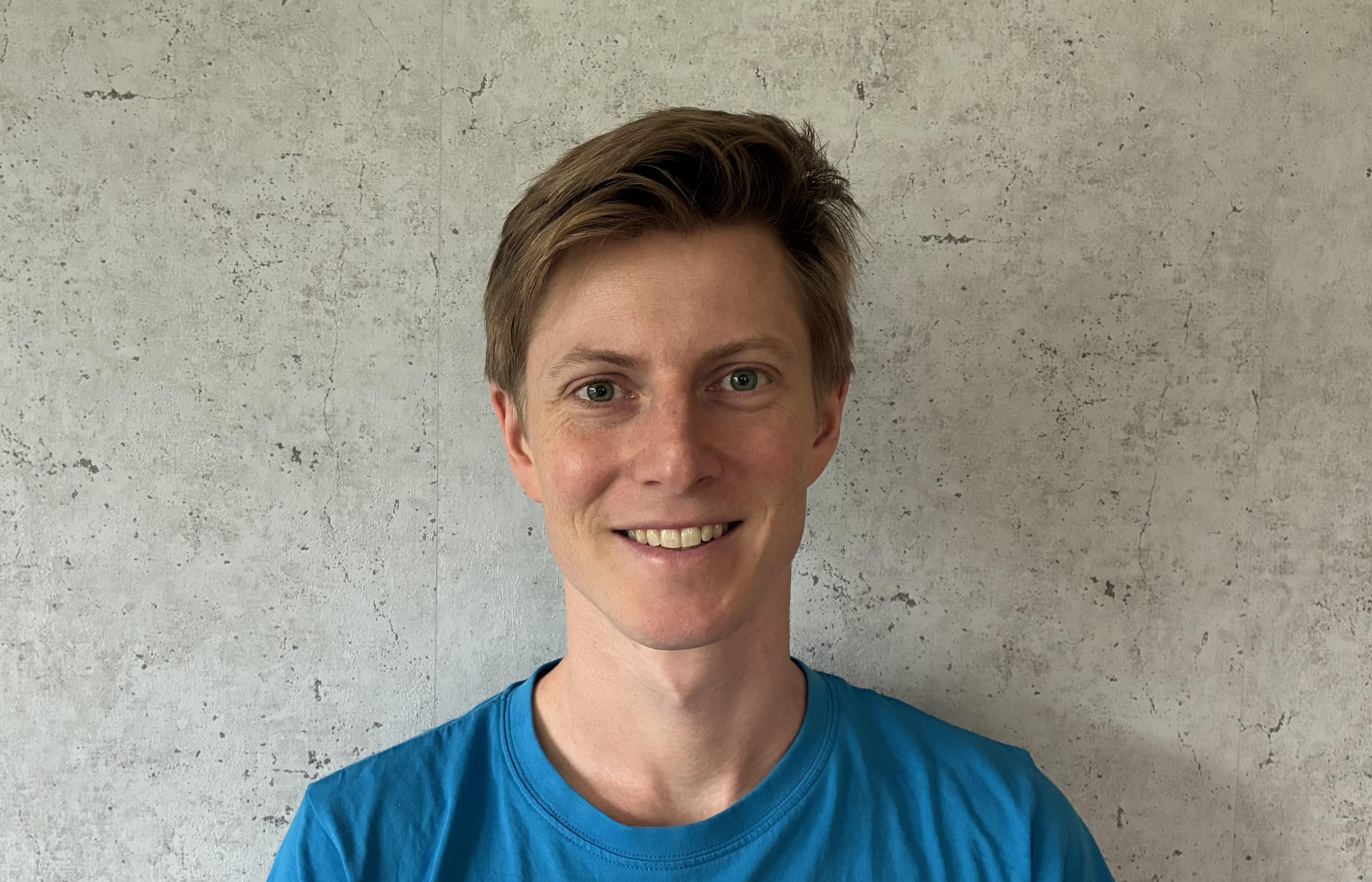A person with short light brown hair wearing a blue shirt, smiling at the camera against a gray textured wall.