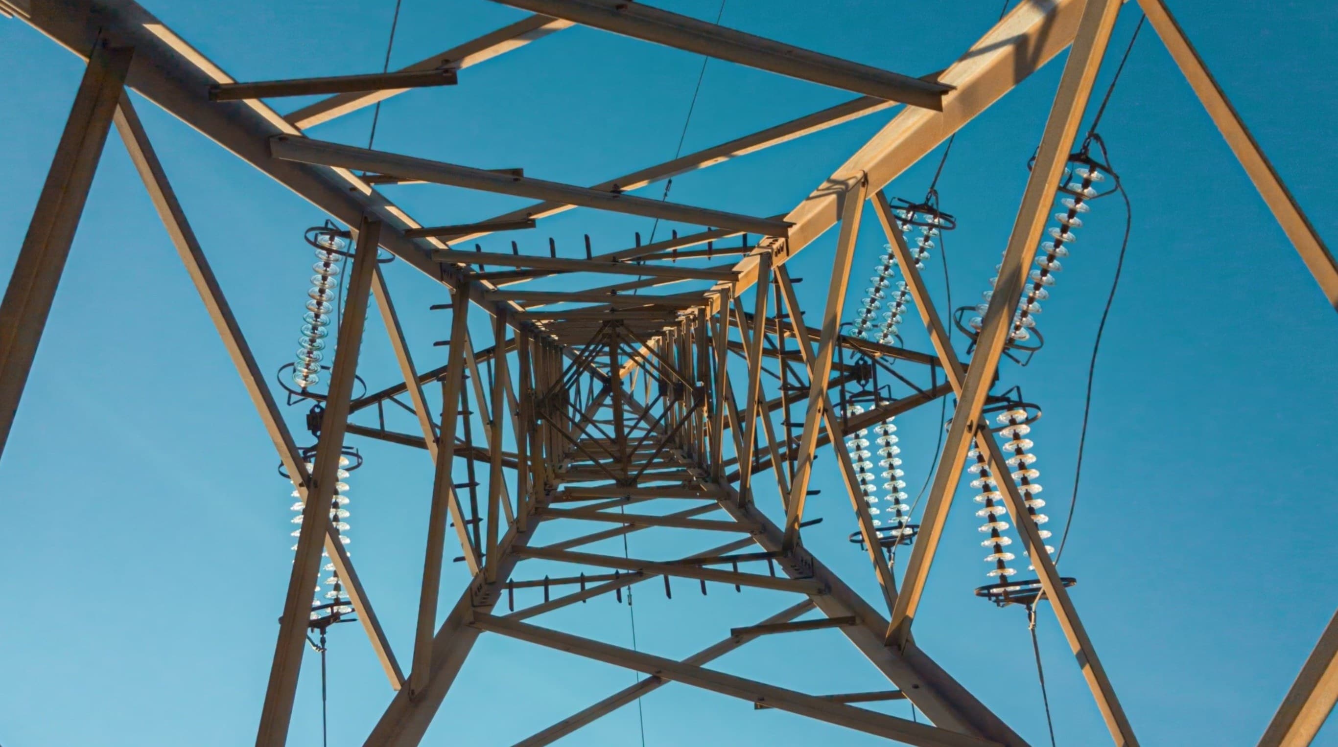Viewed from below, a tall metal transmission tower with crisscrossing beams and hanging insulators, set against a clear blue sky.