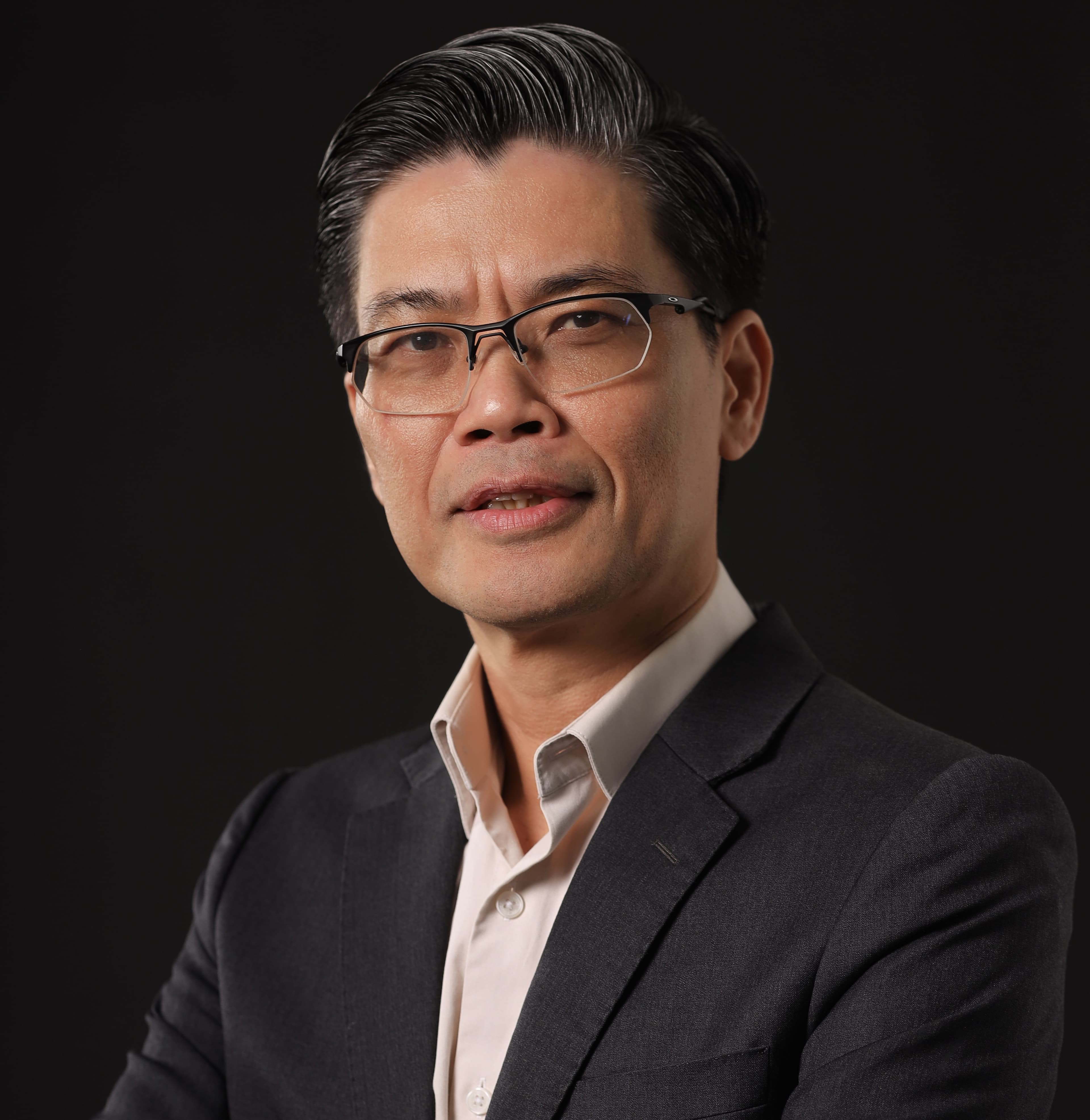 Portrait of a person with short dark hair and glasses, wearing a dark suit and light shirt, against a black background.