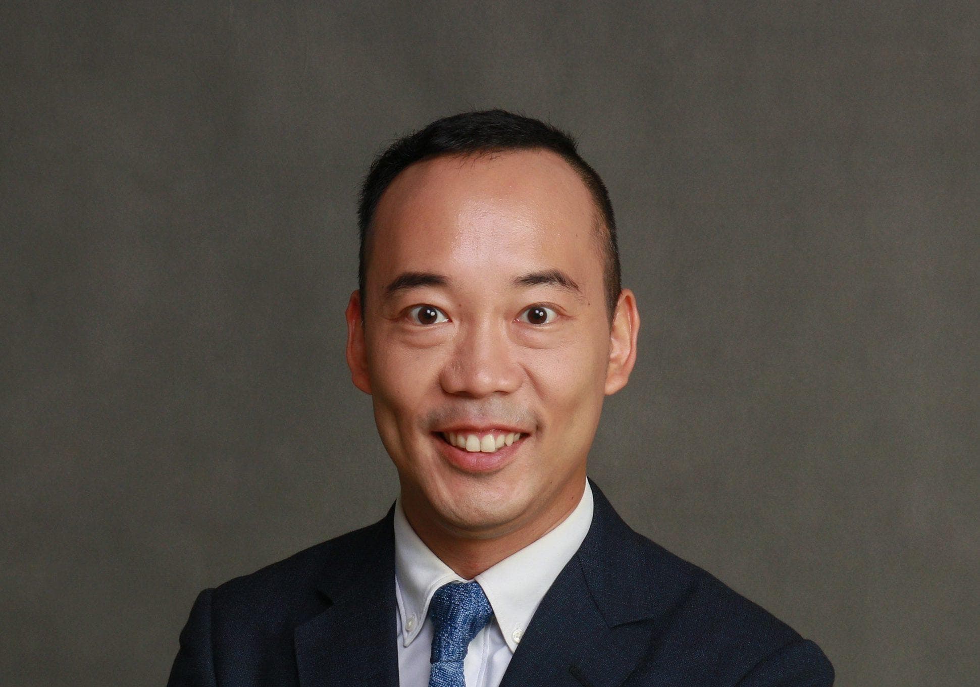 A person in a dark suit, white shirt, and blue tie smiles at the camera against a neutral gray studio backdrop.