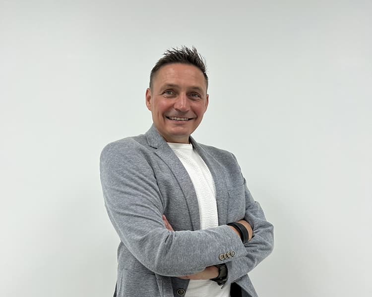 A person with short dark hair, wearing a light gray blazer over a white shirt, stands with arms crossed and smiles against a plain white background.
