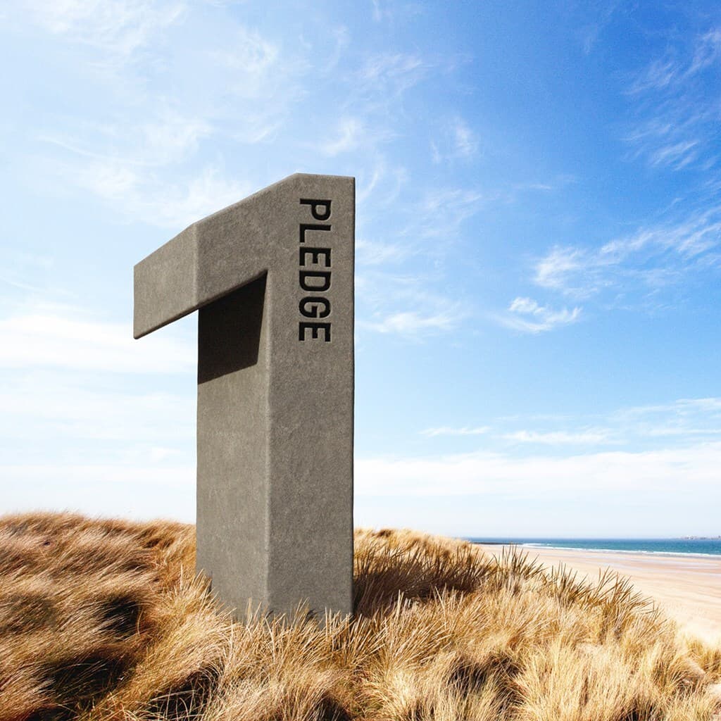 Tall gray concrete sculpture with a horizontal arm rises from dune grasses on a sandy beach, with blue sea behind and the word PLEDGE vertical on its side.
