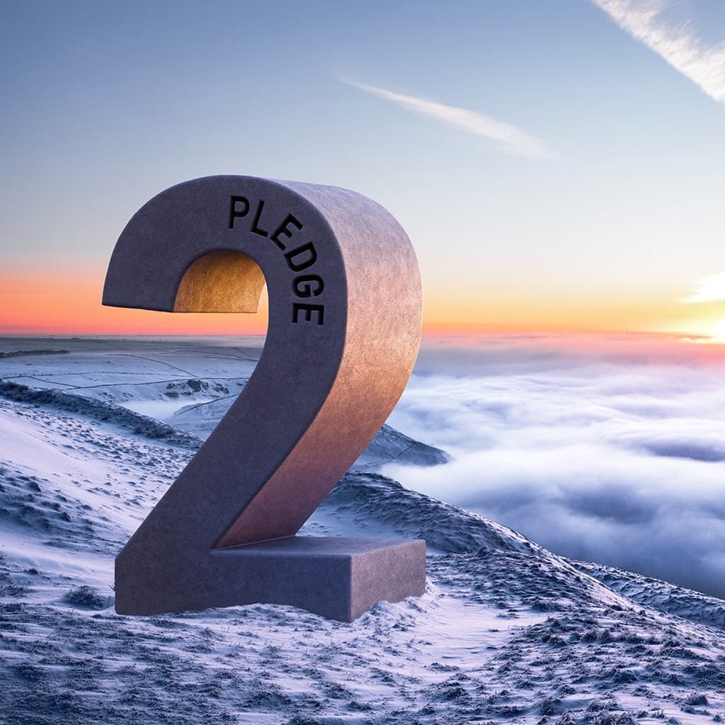 Large curved stone sculpture shaped like the number 2 with 'PLEDGE' along its curve, on a snow-covered mountain ridge at sunrise above clouds.