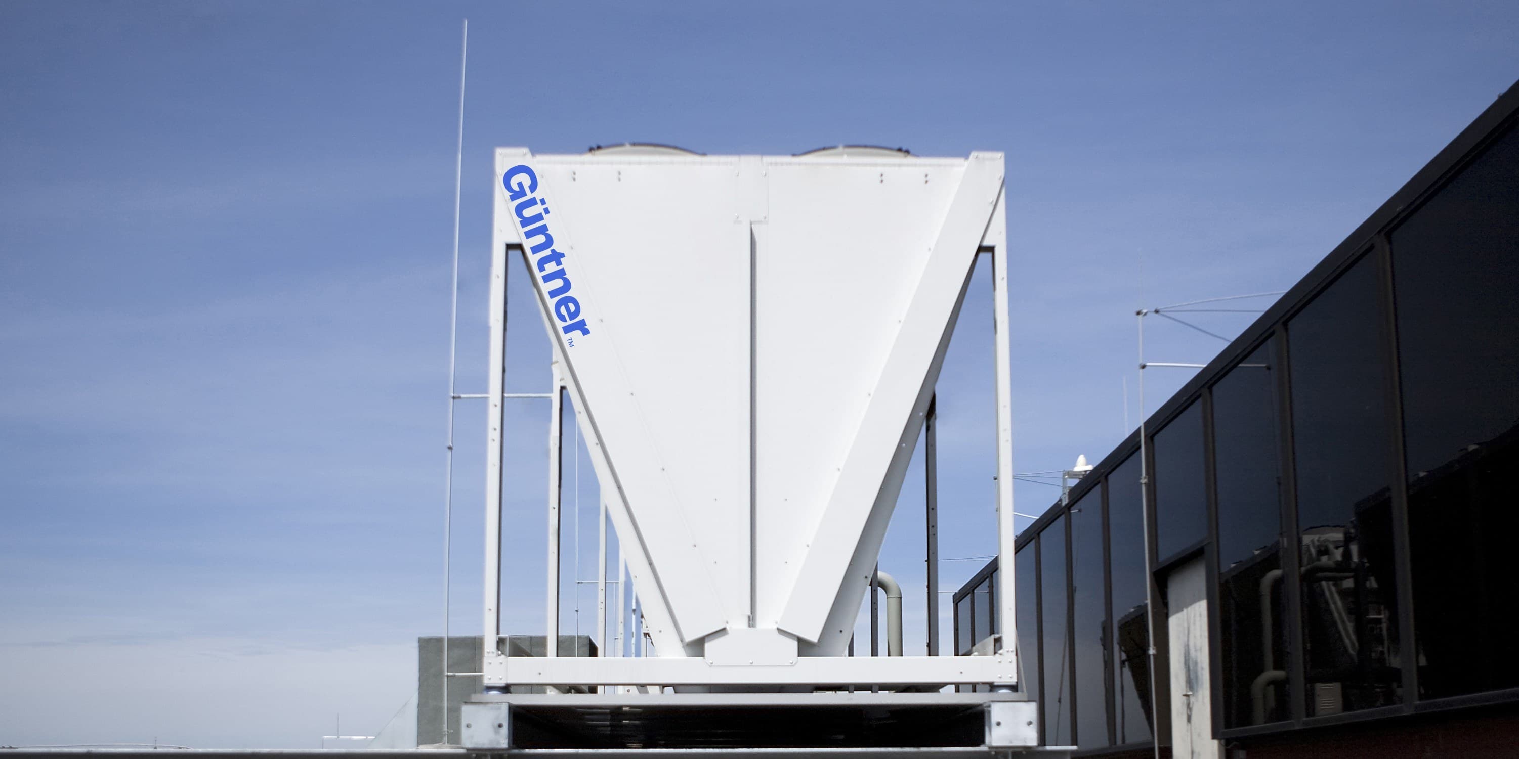 Large white industrial hopper on a rooftop, with a blue Günther logo along its left edge; glass-walled building to the right under a clear blue sky.