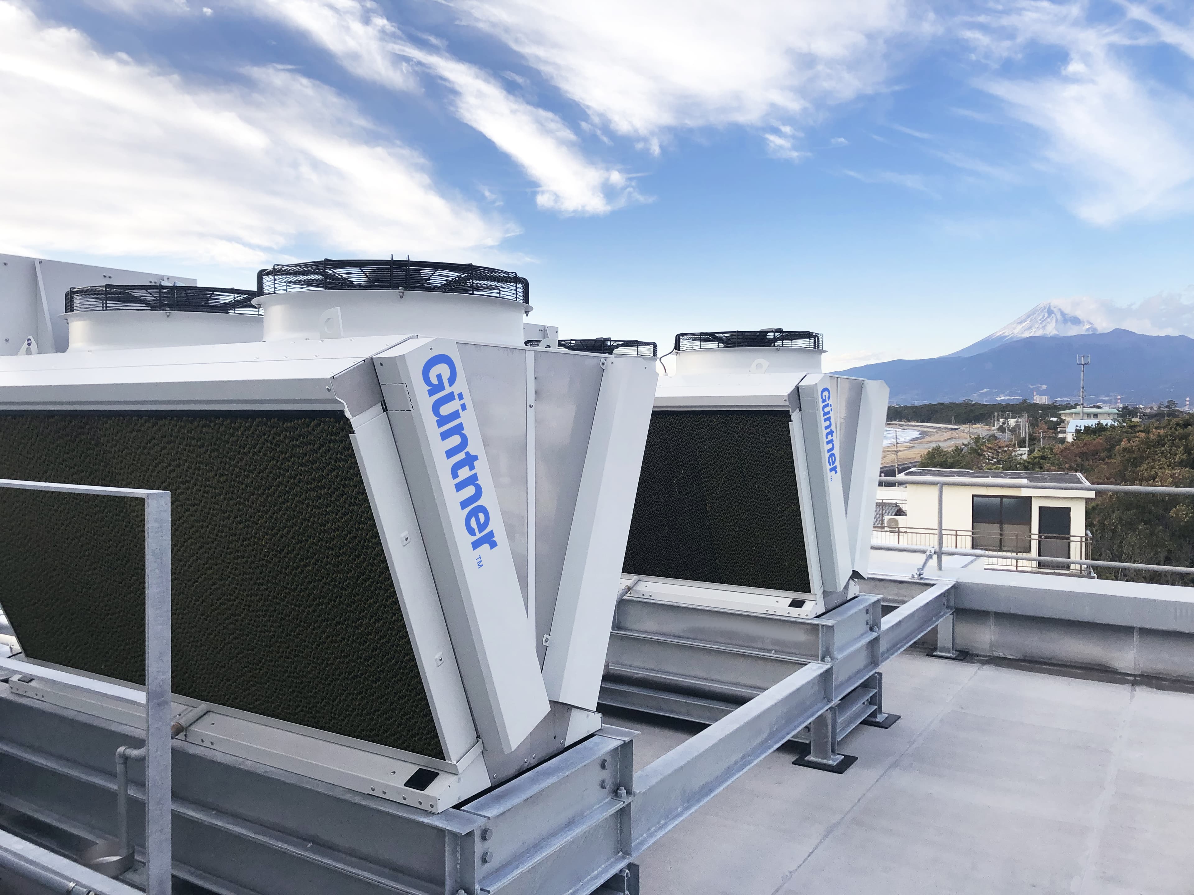 Rooftop HVAC units with white frames, dark fins and circular top fans, labeled Guntner, mounted on a metal platform, blue sky with wispy clouds and a distant snow-capped mountain.