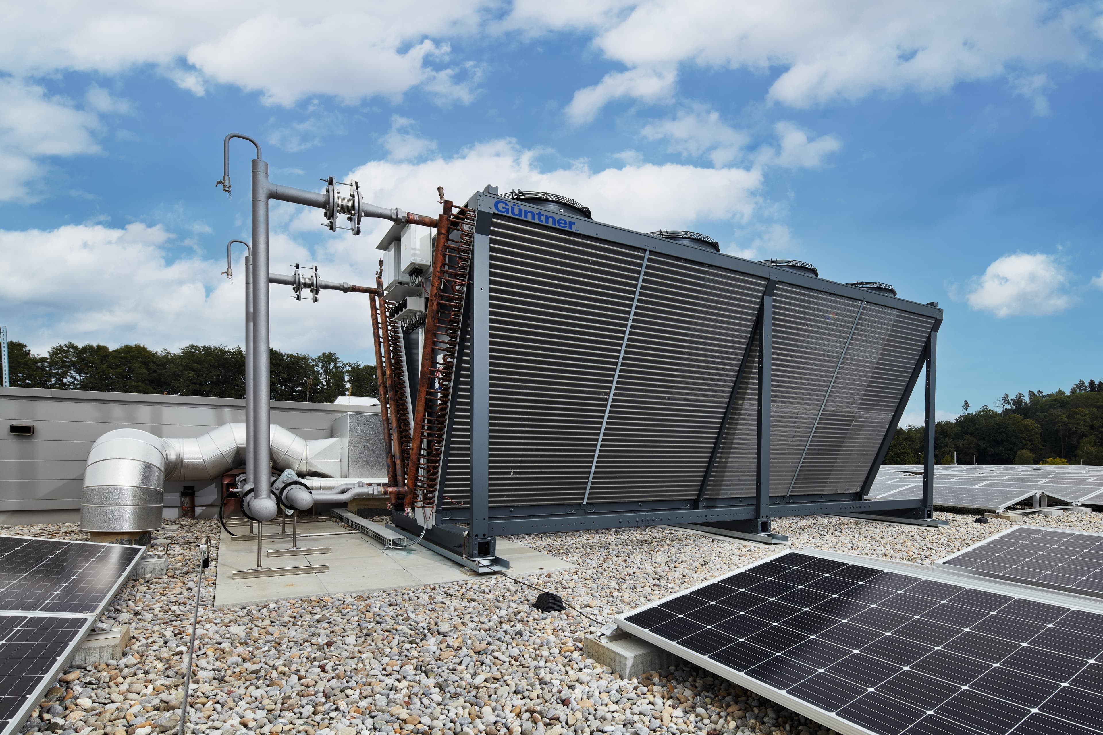Rooftop scene with a large ribbed metal cooling unit labeled Guntner, connected pipes, and solar panels under a blue sky with scattered clouds.