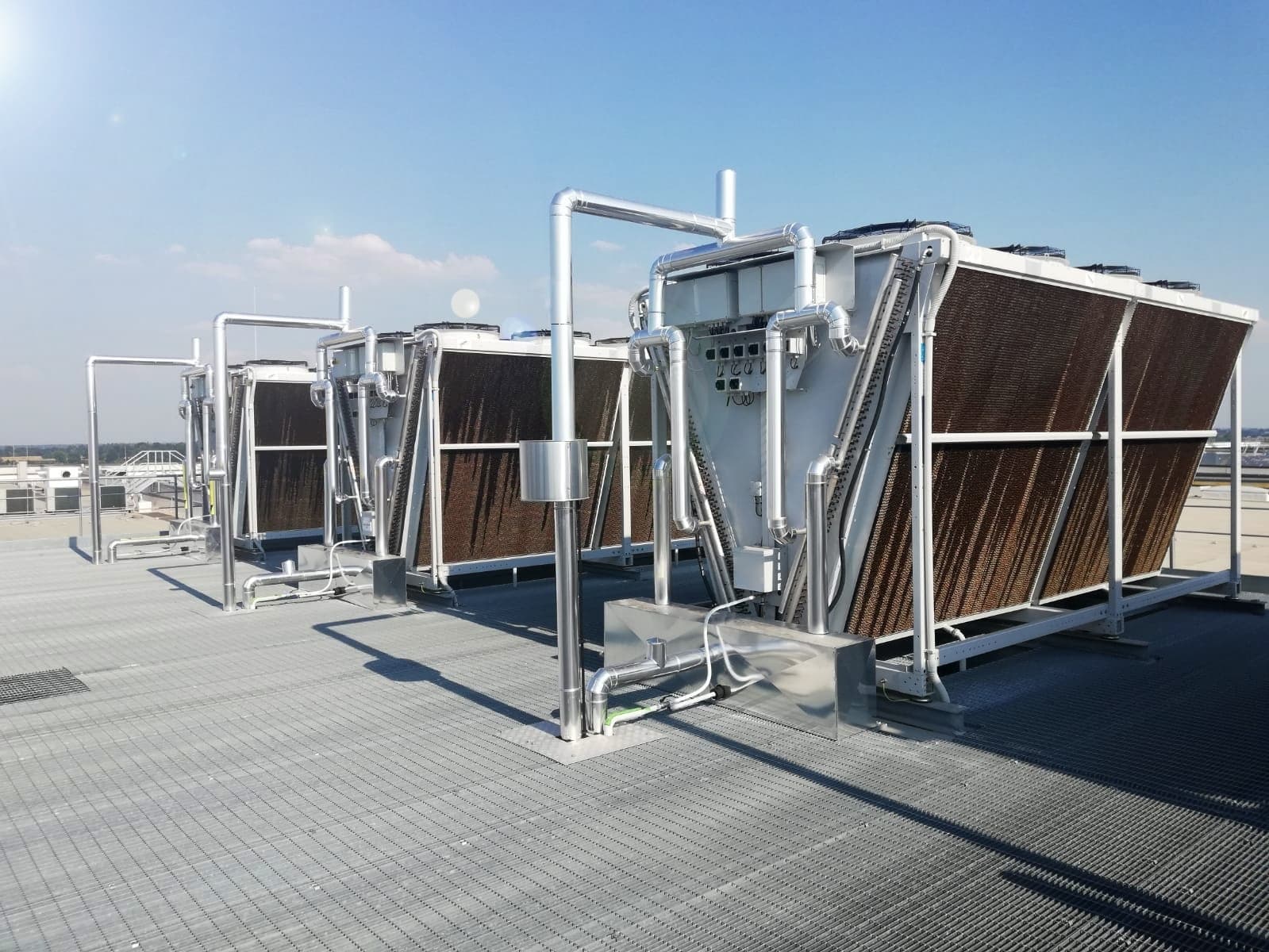 Rooftop with large industrial heat exchangers and cooling units, silver piping, and metal framing on grated decking under a clear blue sky.