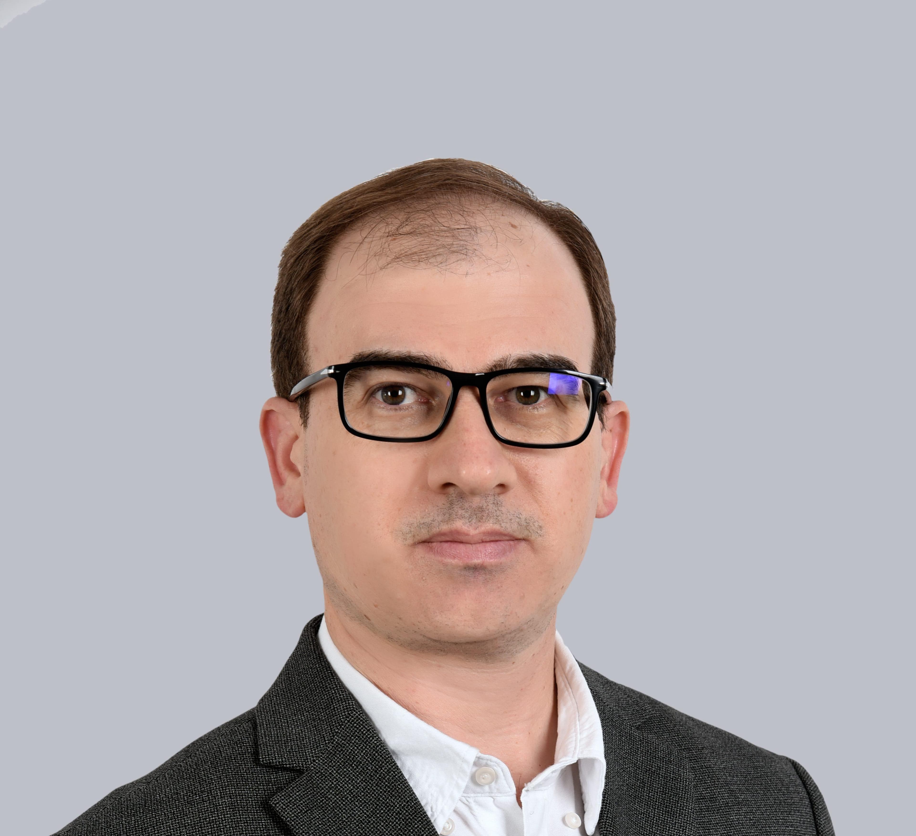 Portrait of a person with short brown hair and black-framed glasses, wearing a dark blazer and white shirt, against a plain light-gray background.