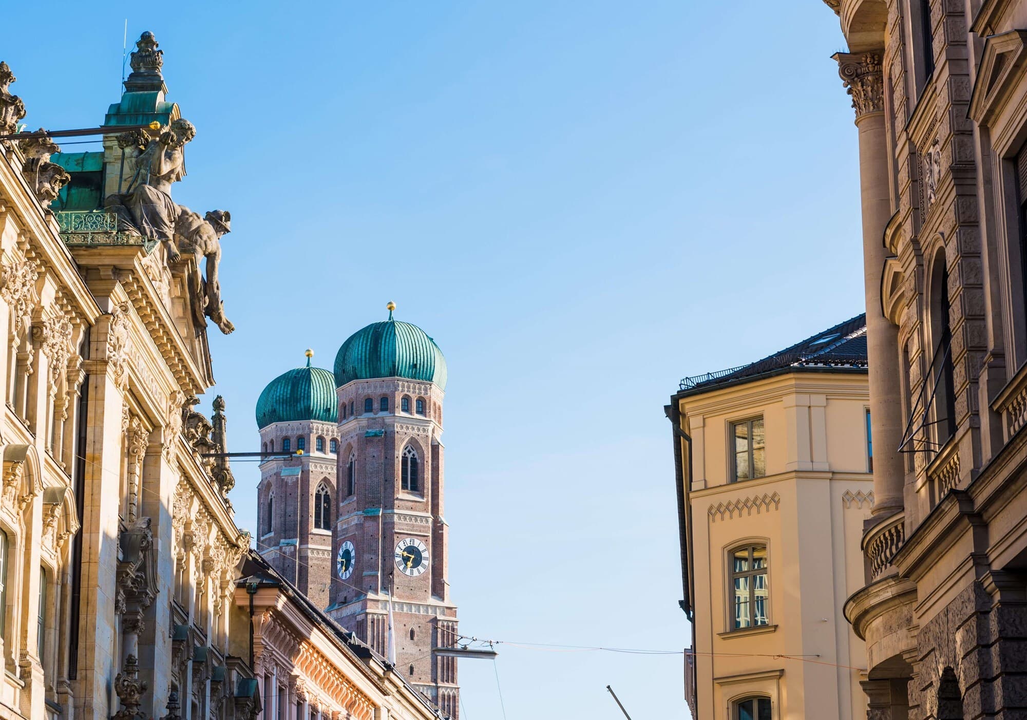 A sunlit European street with ornate historic facades and statues on the left, and twin green-domed church towers above brick buildings against a clear blue sky.