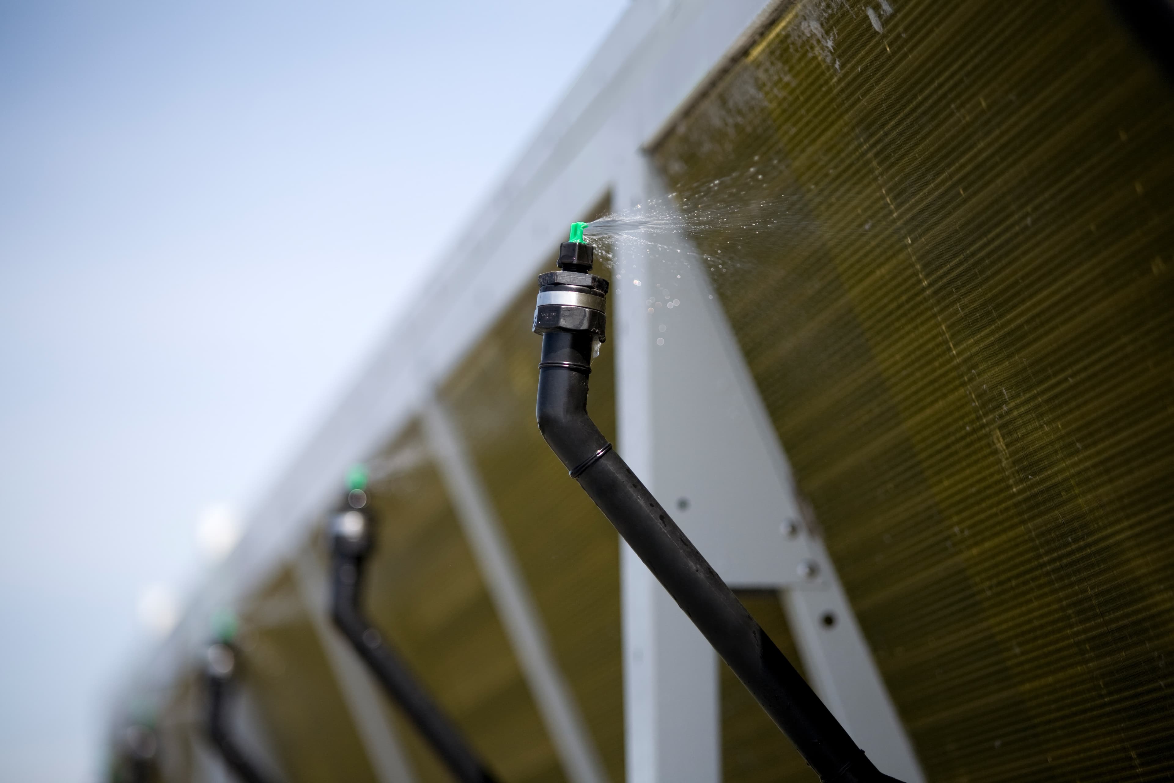 Duză verde a unui furtun pulverizează apă pe panouri solare montate pe structură metalică; imagine de aproape, cu panouri în fundal.