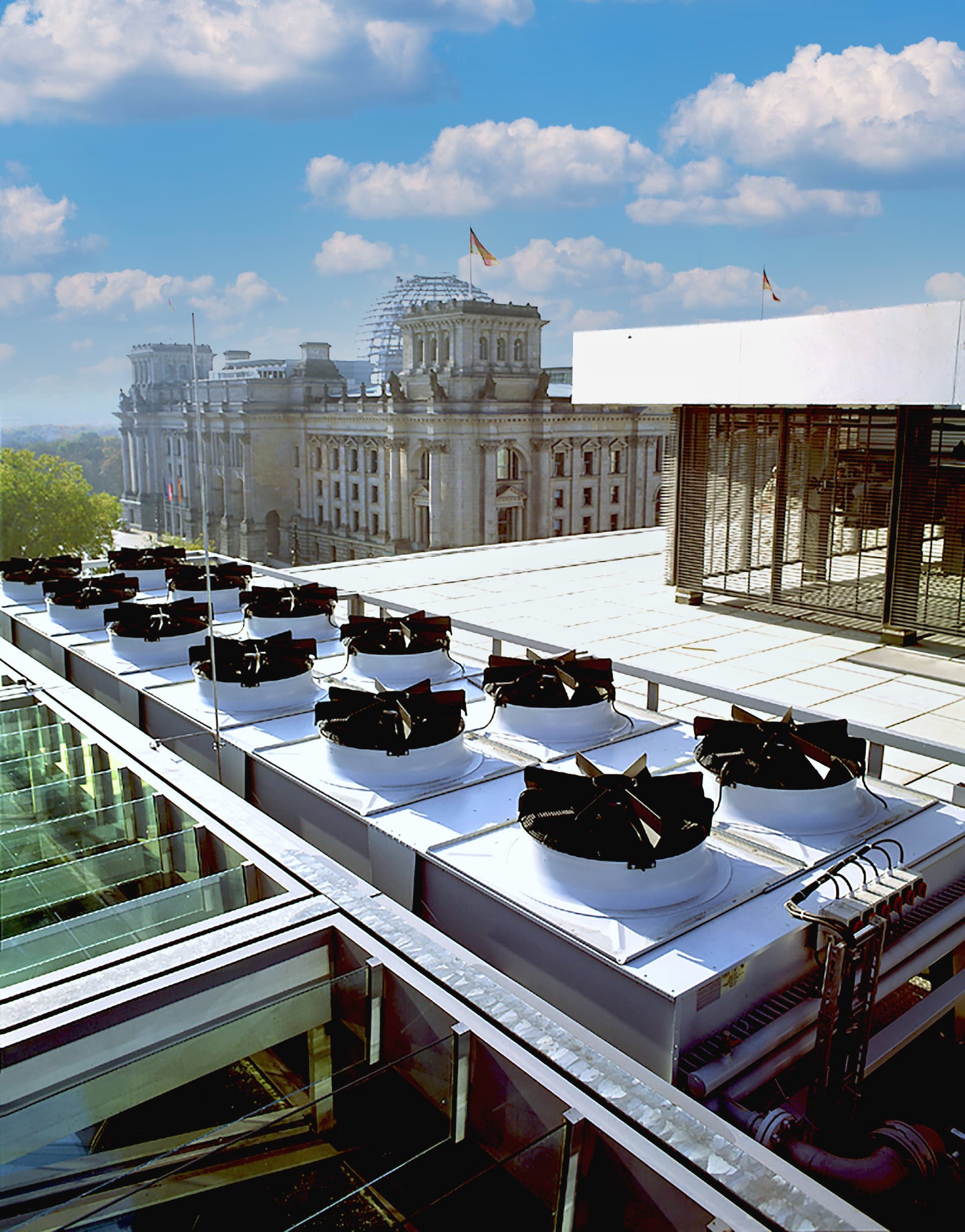 Rooftop with multiple circular cooling fans and pipes; a grand historic building with flags and a glass dome in the background under a blue sky.