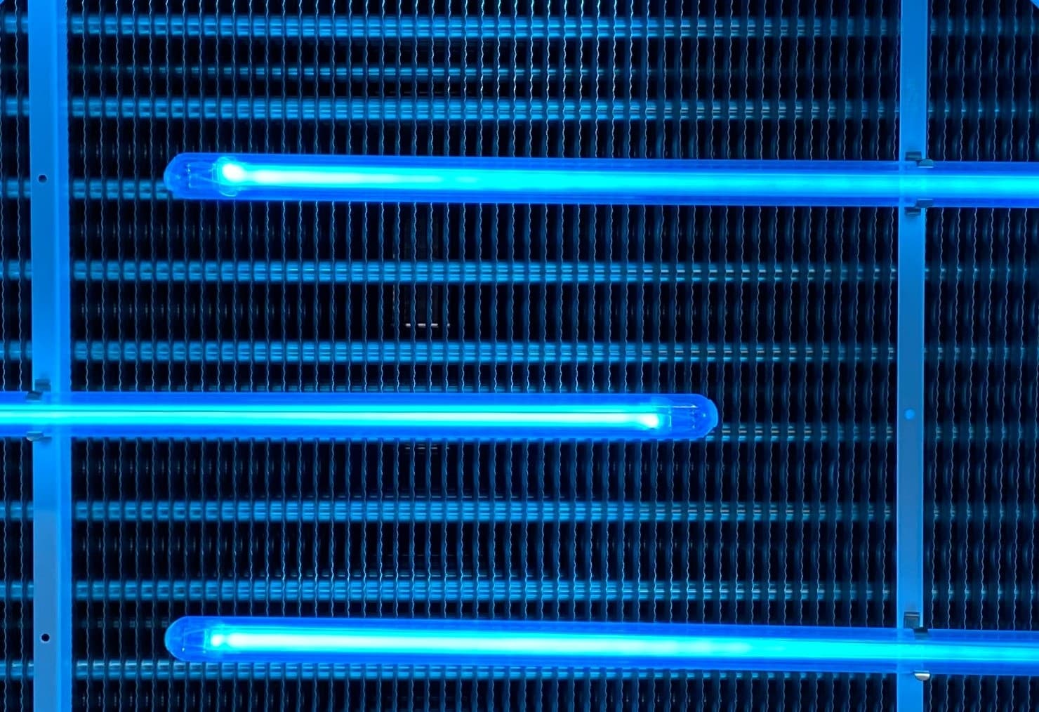 Three blue neon fluorescent tubes mounted on a metal frame glow over a dark ribbed background.