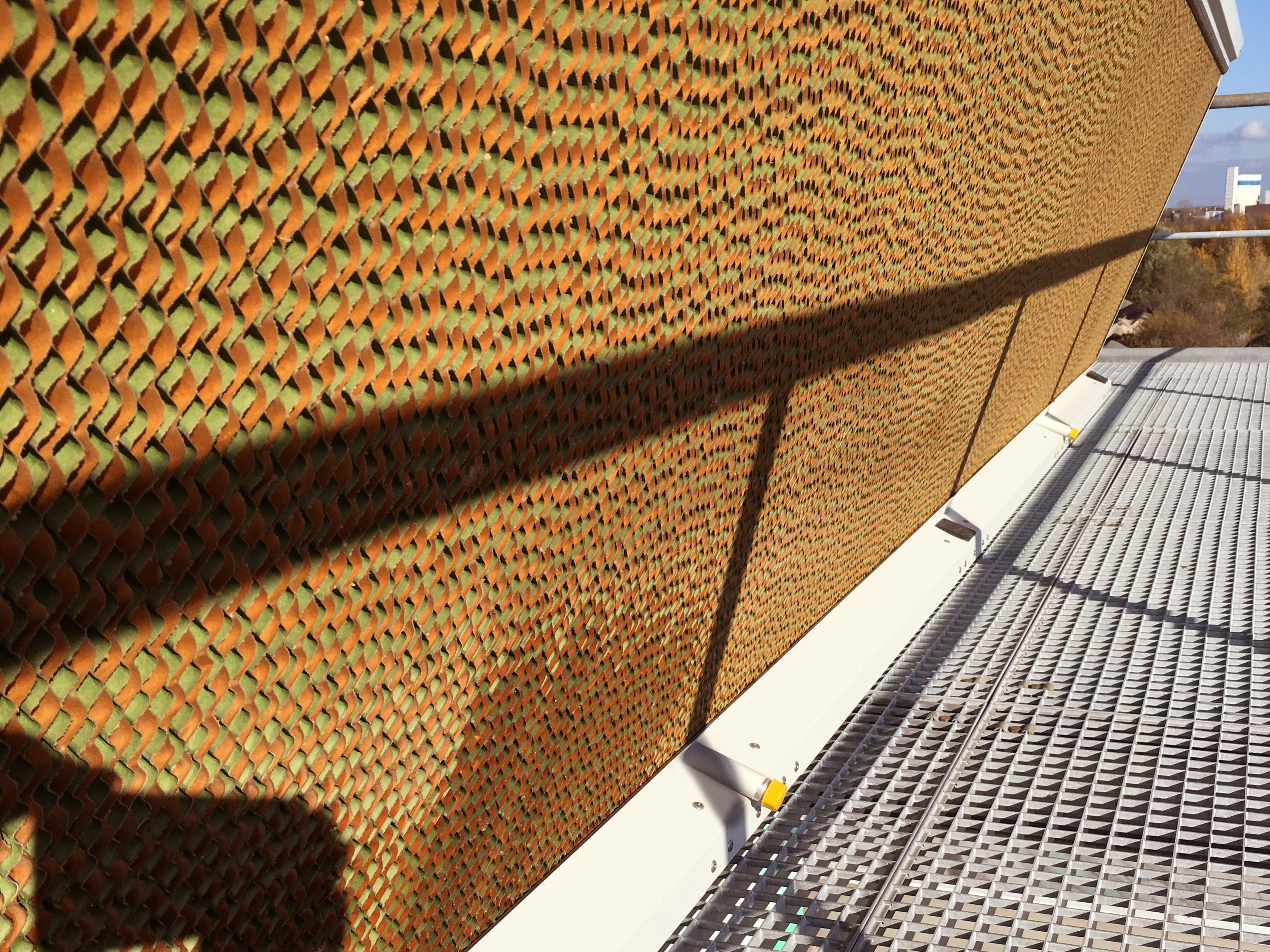 Curved orange perforated lattice wall with a diamond pattern, casting a long shadow across a metal grated rooftop walkway.