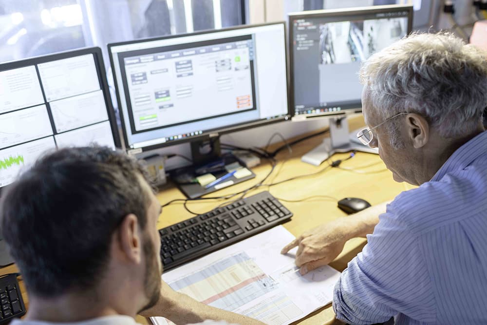 Two people sit at a desk with multiple computer monitors displaying dashboards, while one points at a large printed plan.