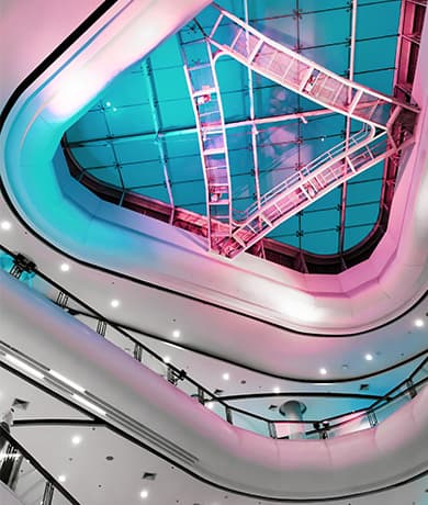 Interior atrium of a multi-level mall with curved white balconies, a glass skylight, and neon pink–blue lighting.