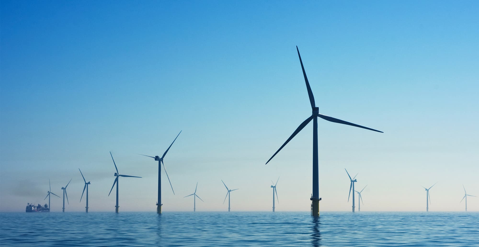 Offshore wind farm with tall turbines rising from calm blue sea under a clear sky; a distant ship is visible near the horizon on the left.