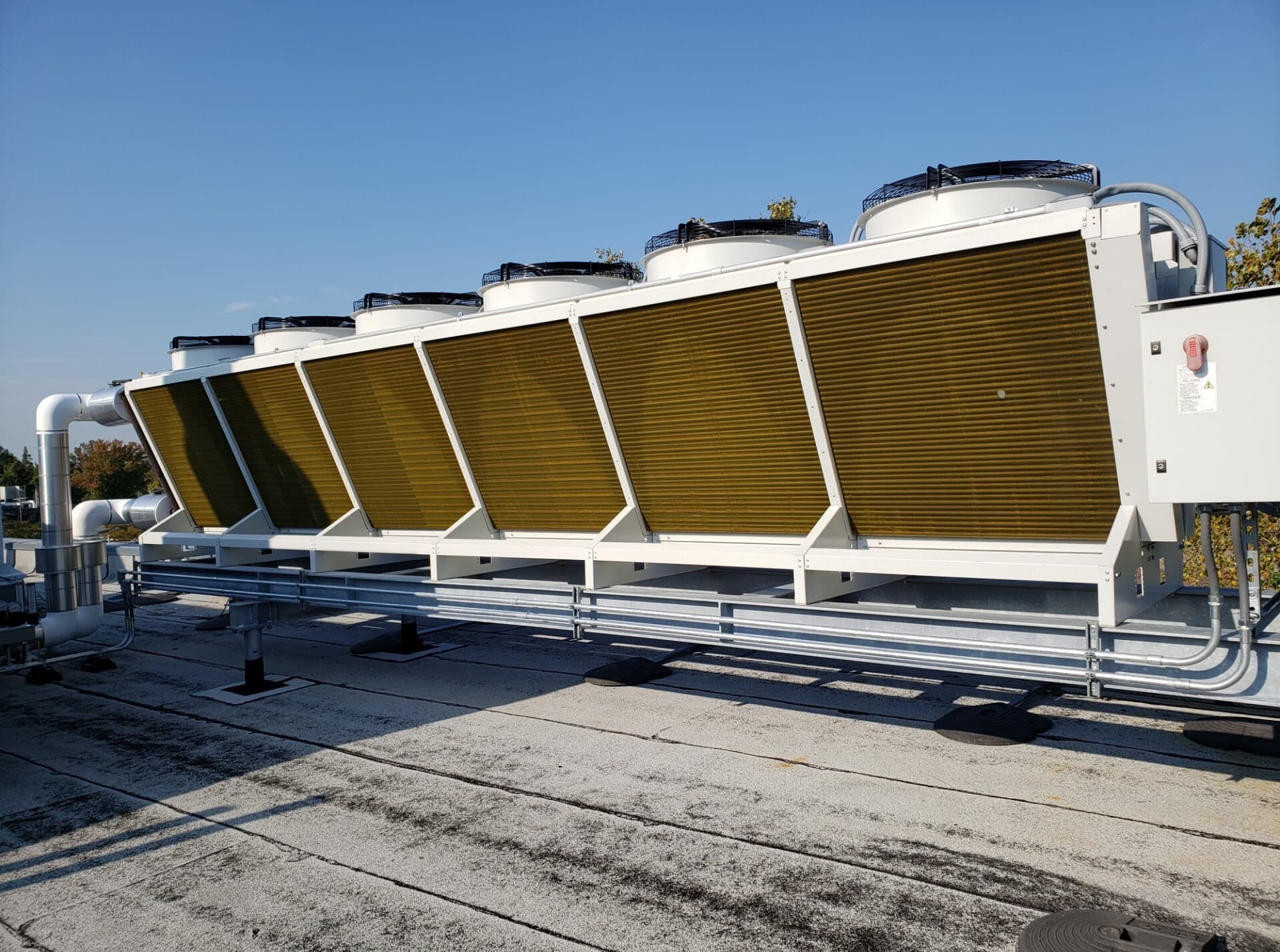 Rooftop industrial cooling units with angled brown fins and large circular fans, mounted on white frames with exposed pipes under a clear blue sky.