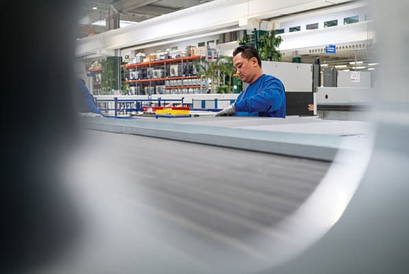 A person in a blue shirt works at a factory workstation, inspecting items on a conveyor, with shelves and plants visible in the background.