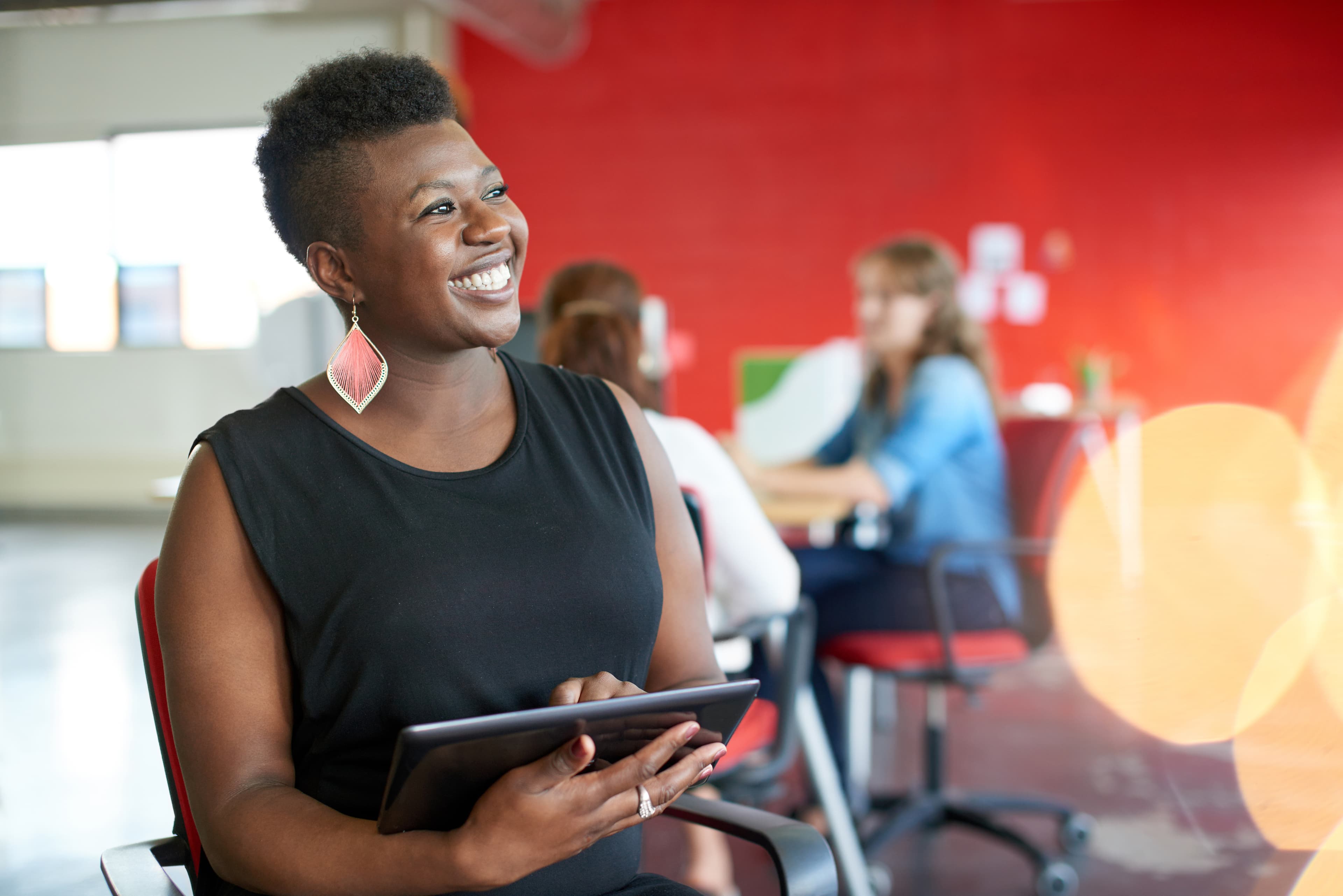 Person with short hair, wearing a black sleeveless top and pink teardrop earrings, smiling while holding a tablet in a bright office with a red wall.