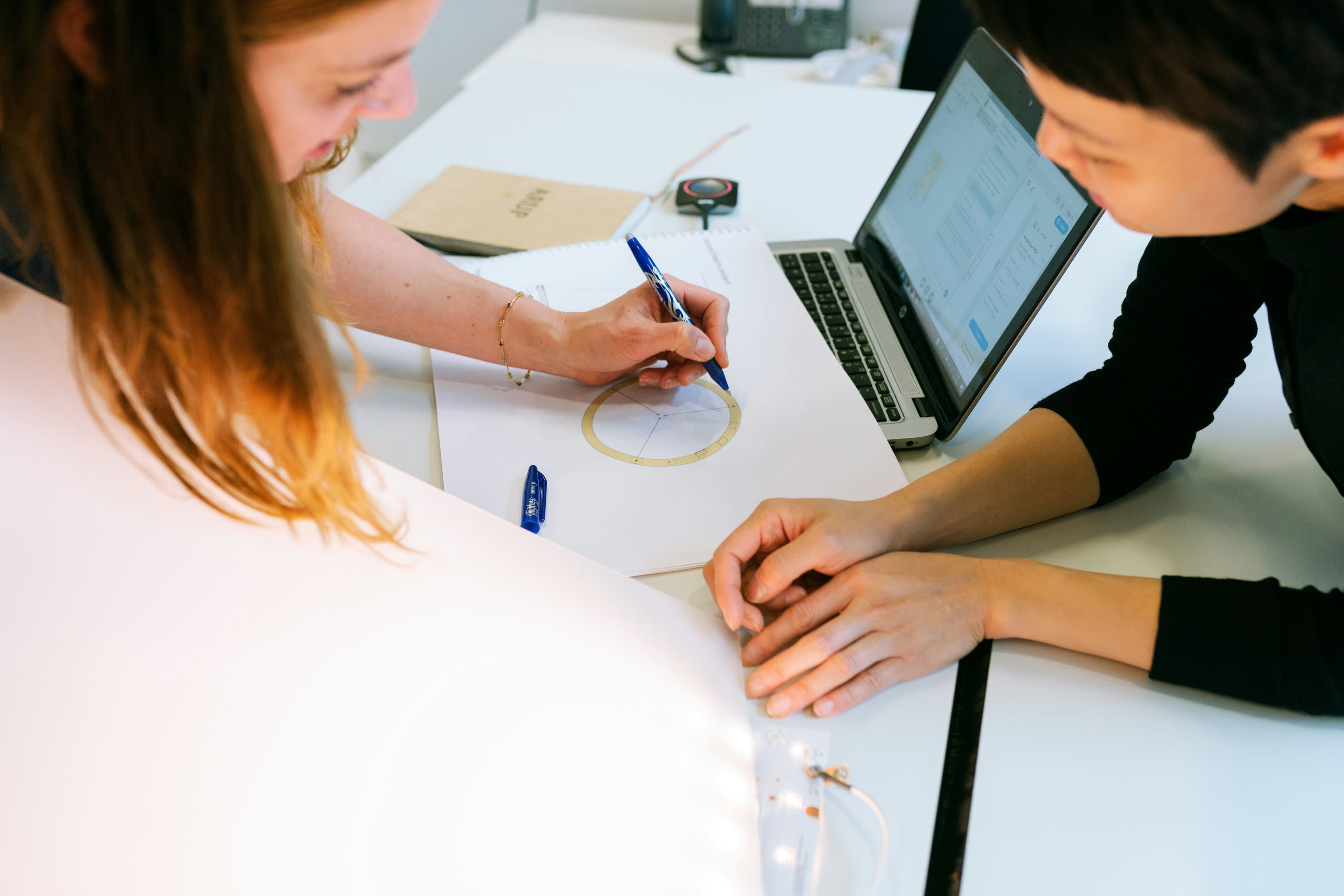 Two people collaborate at a white desk; one draws a circle with a blue pen while the other looks at a laptop, with markers and a notebook nearby.