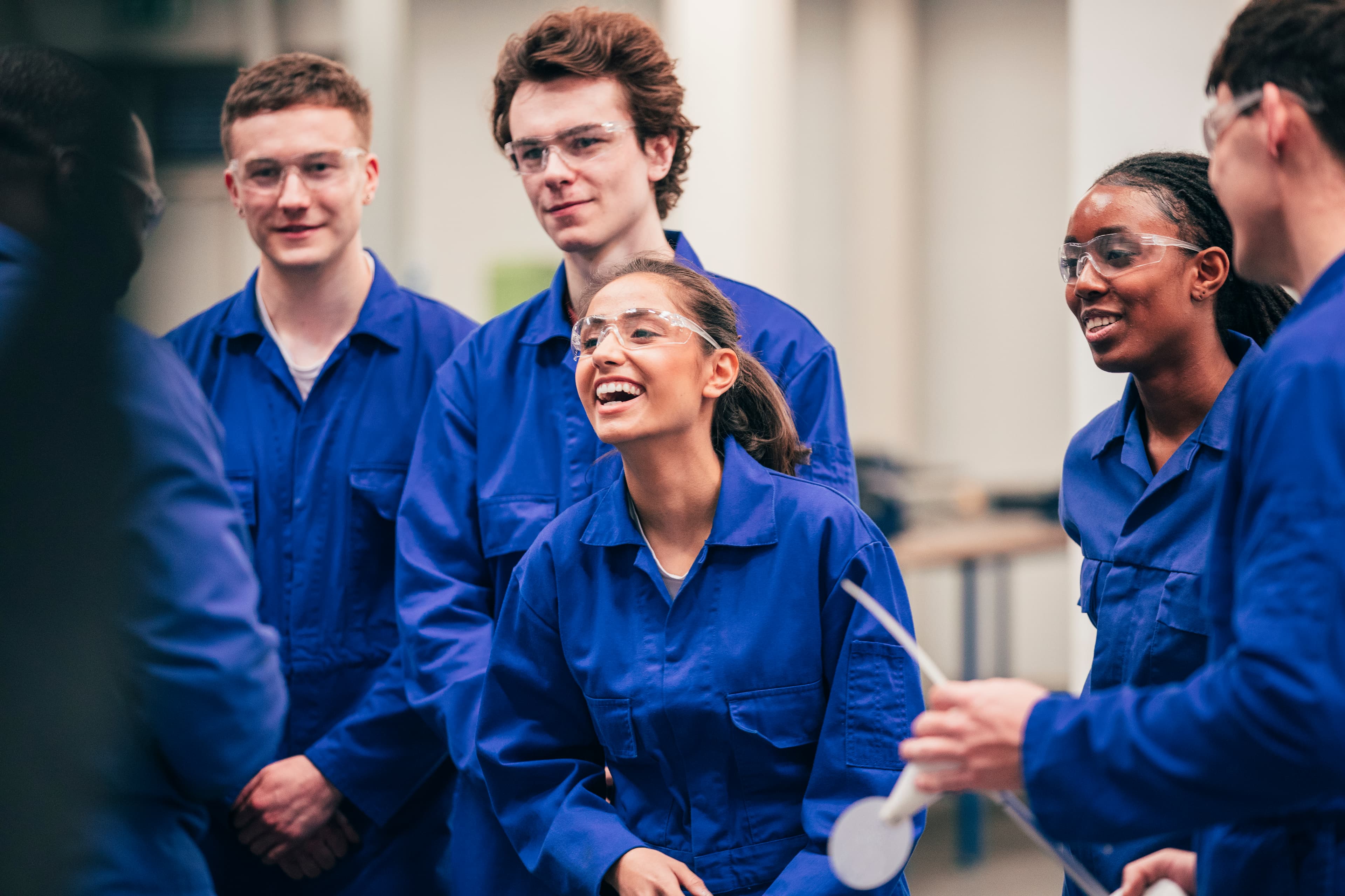Group of students in blue coveralls and safety goggles, smiling and chatting in a lab or workshop environment with equipment in the background.