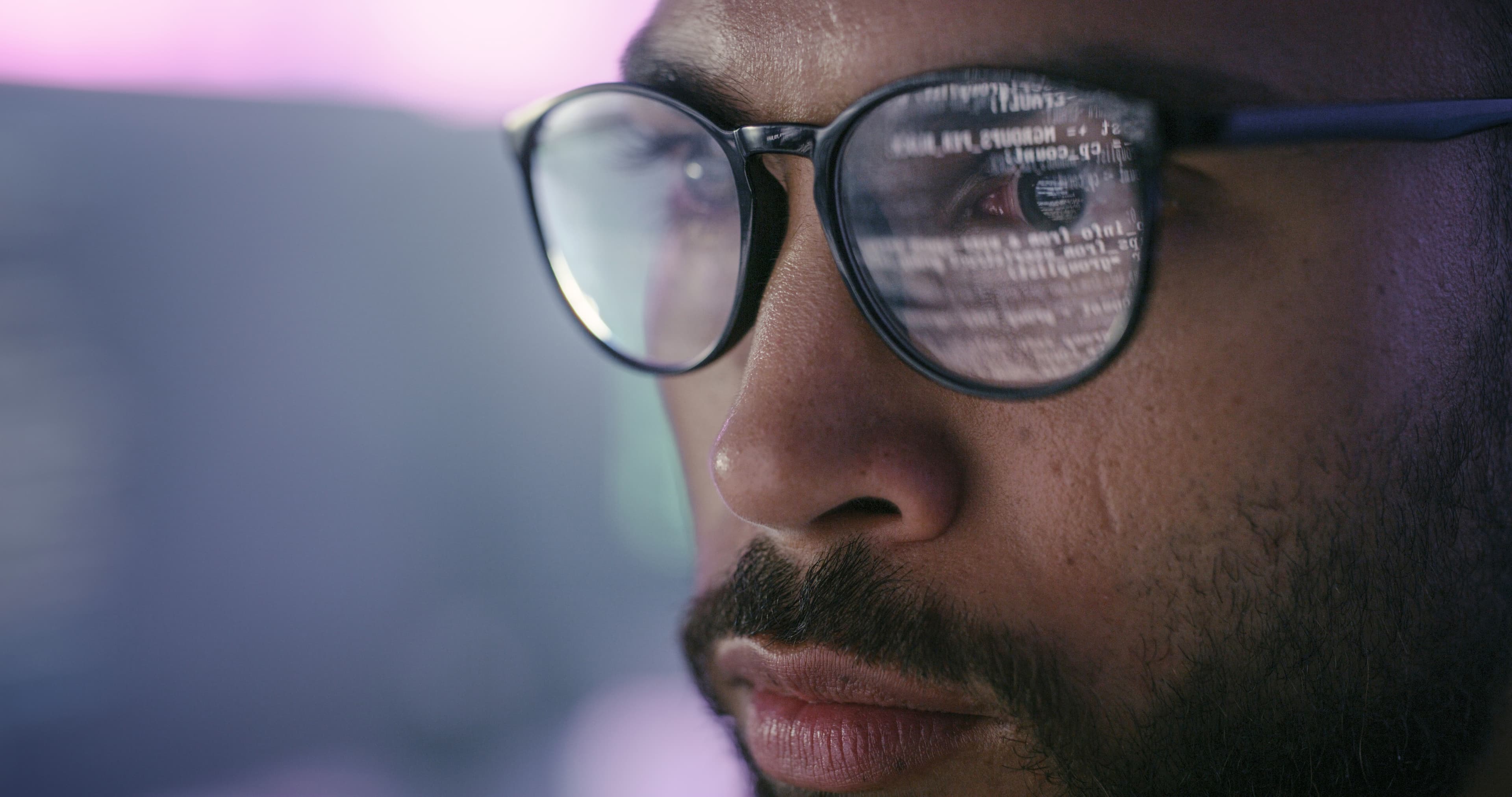 Close-up of a person with a beard wearing glasses; code reflected in the lenses, with a soft purple-blue background.