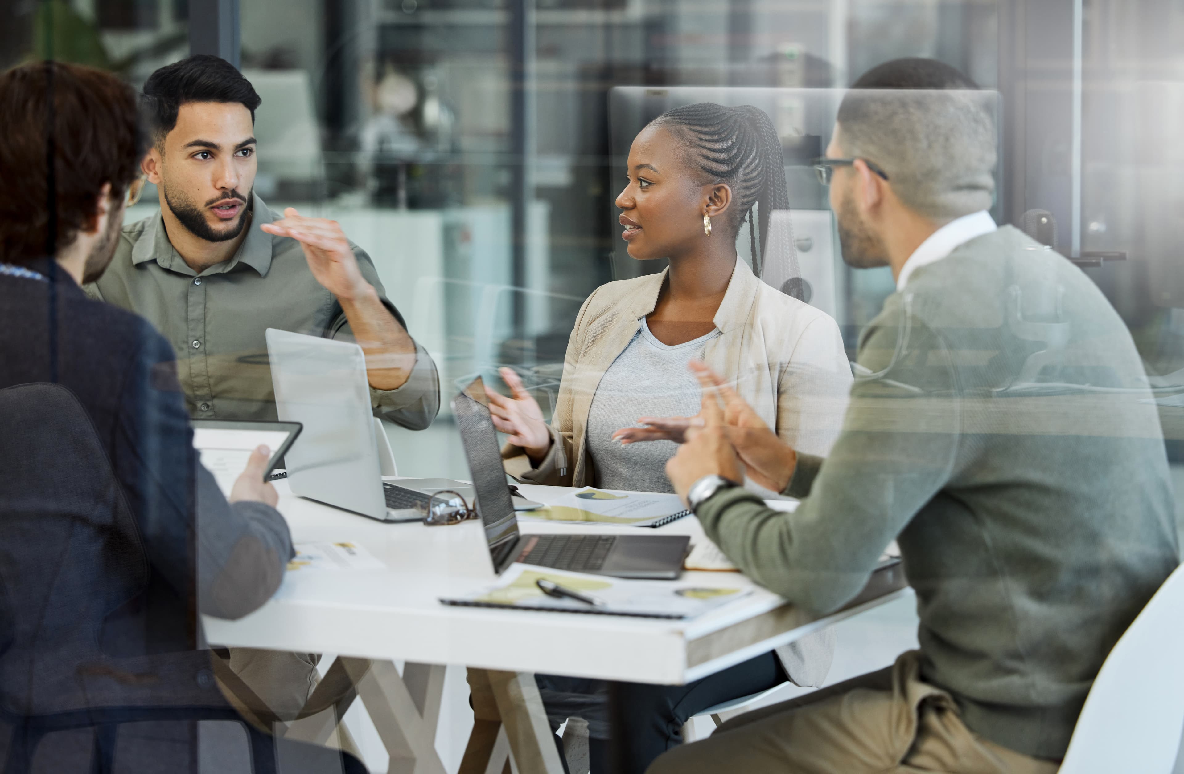 Four diverse coworkers in a glass-walled office sit around a table with laptops and documents, actively discussing with hand gestures.