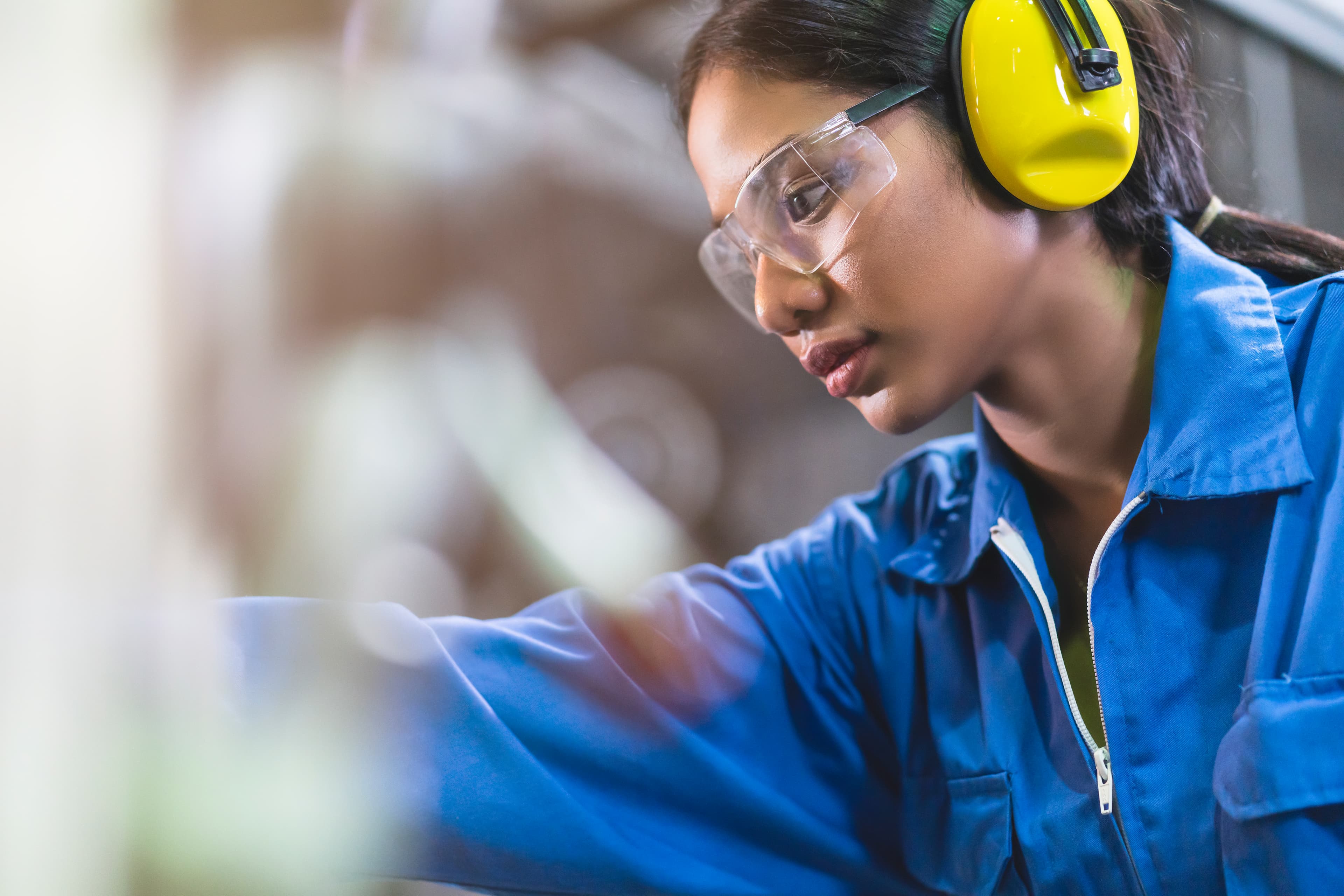 Person in blue coveralls wearing clear safety glasses and yellow earmuffs, leaning to inspect something in a workshop.