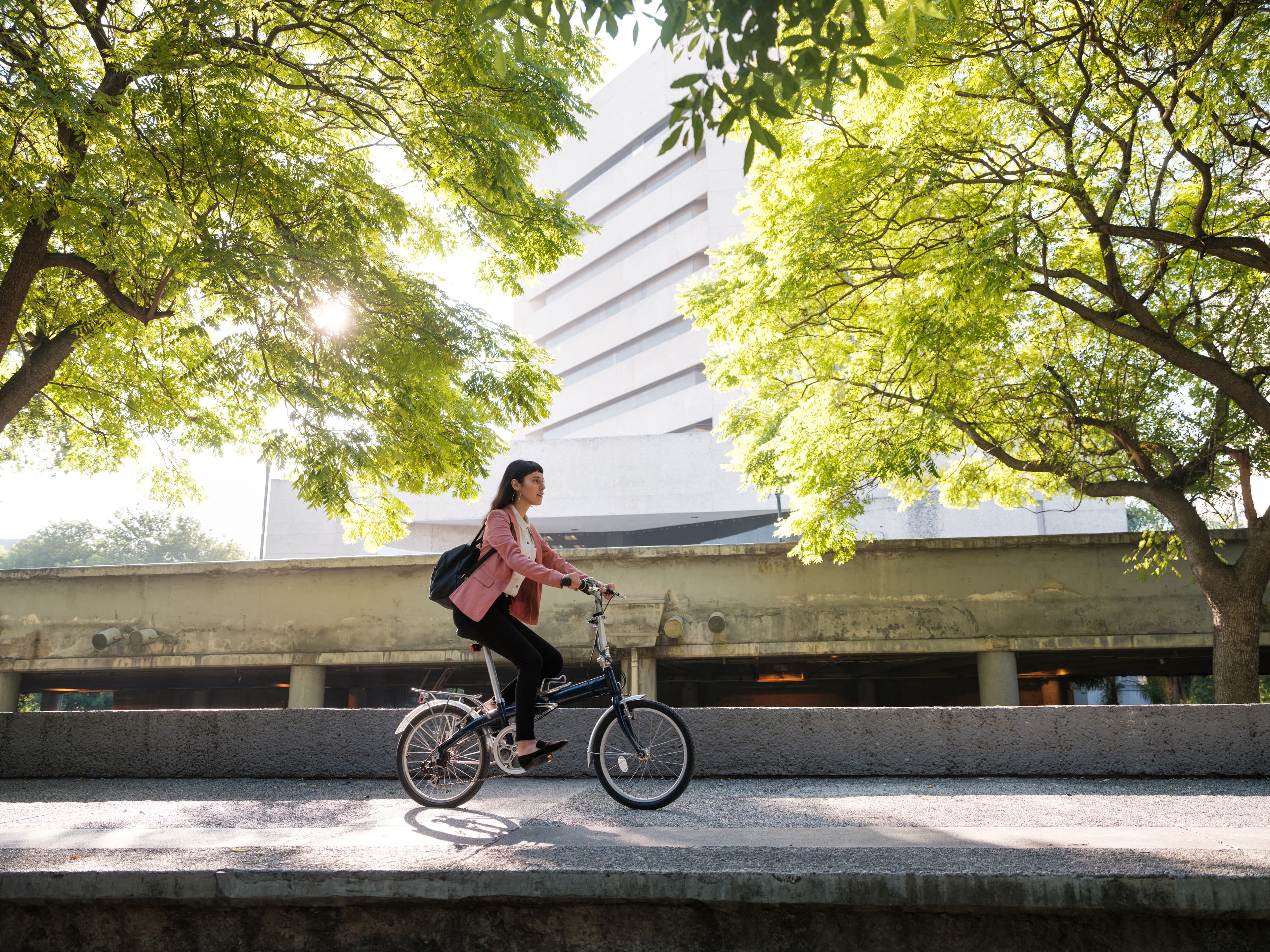 Person in a pink jacket rides a small bicycle on a sunlit street, framed by lush green trees with a concrete building in the background.