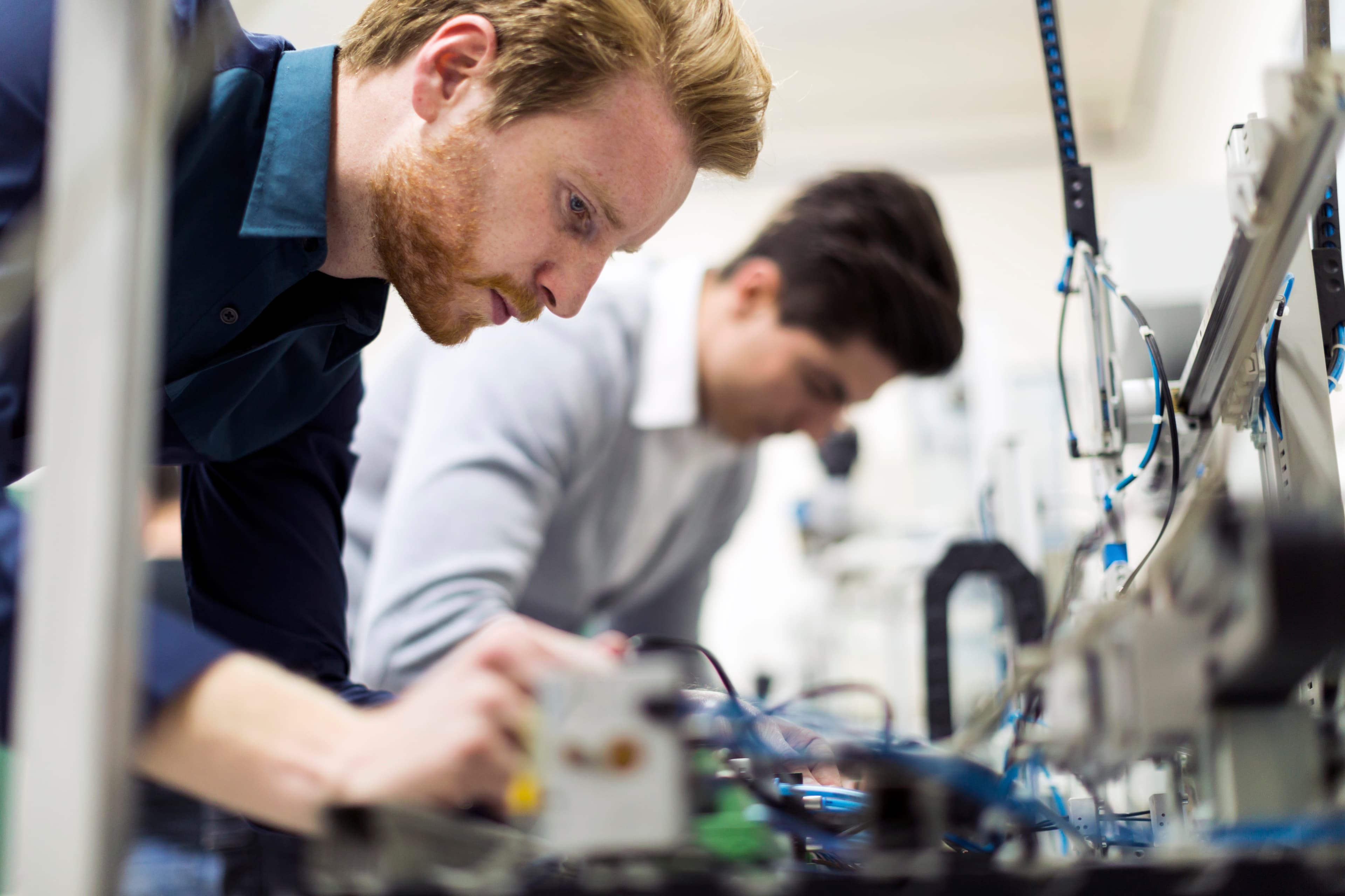 Two individuals work at a lab bench with electronic gear and blue cables; a foreground person leans over a device.