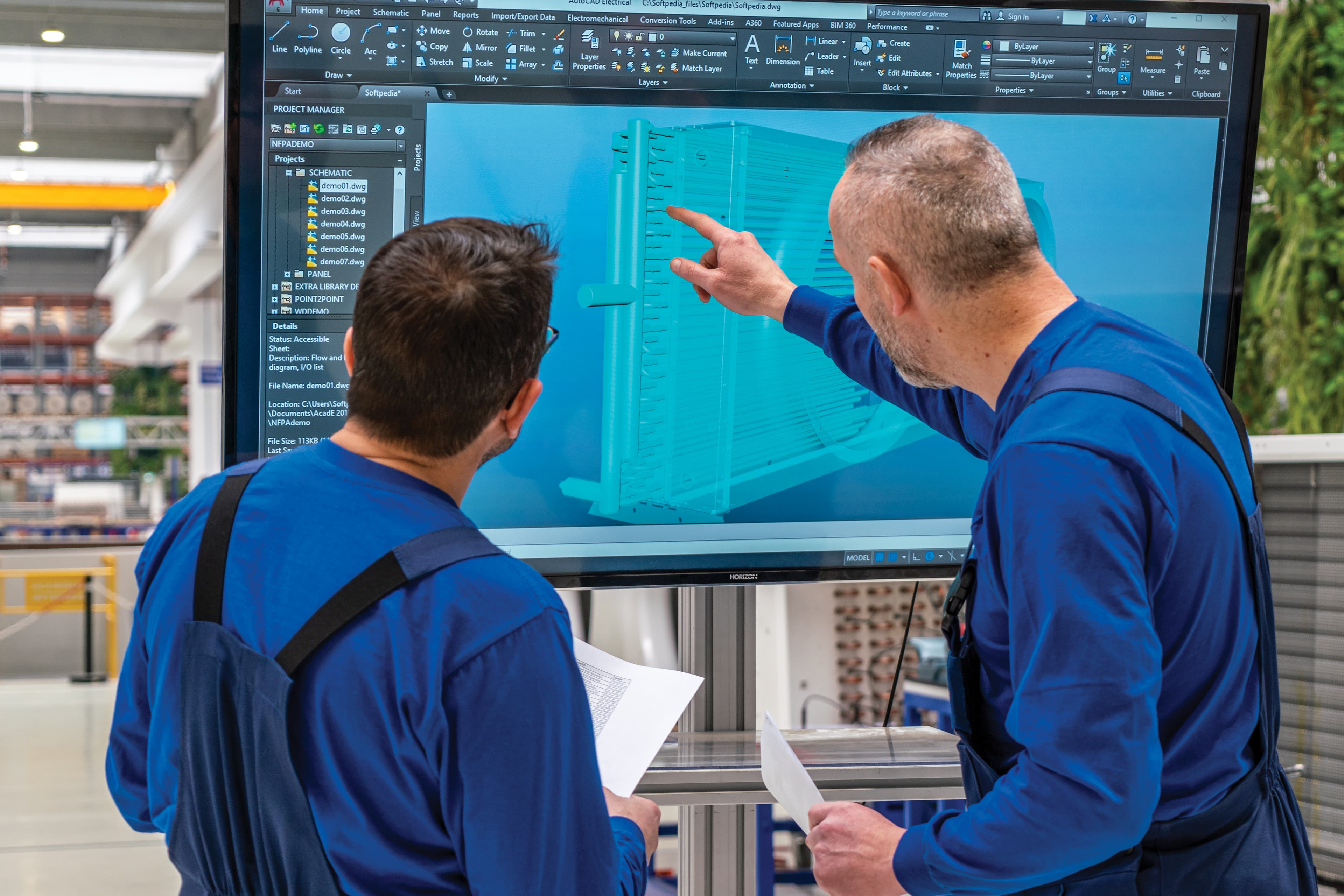 Two workers in blue overalls study a large CAD screen showing a teal 3D model, with one person pointing and both holding papers in an industrial workshop.