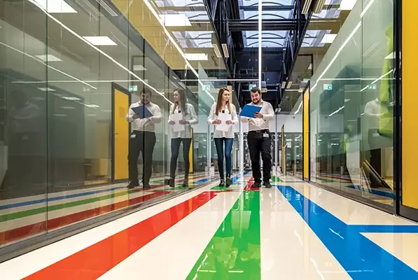 Five people walk down a glass-walled hallway with a bright striped floor, carrying folders and tablets, wearing white tops.