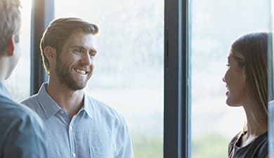 Three colleagues by large windows in a bright office; a person in a light blue shirt smiles while speaking with two others.