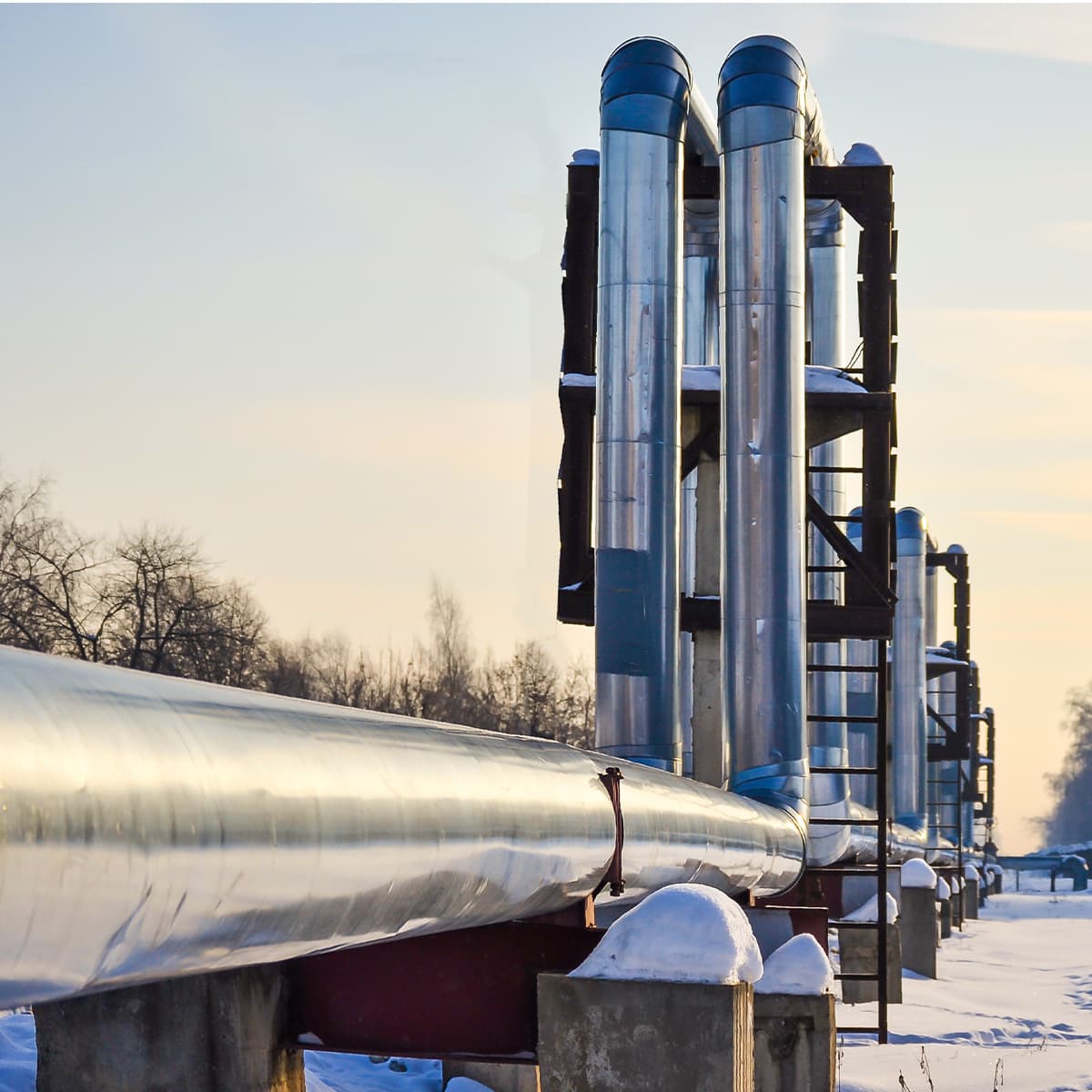 Row of shiny vertical metal pipes on a platform, connected to a long horizontal pipe, in a snow-covered industrial site under a pale winter sky.