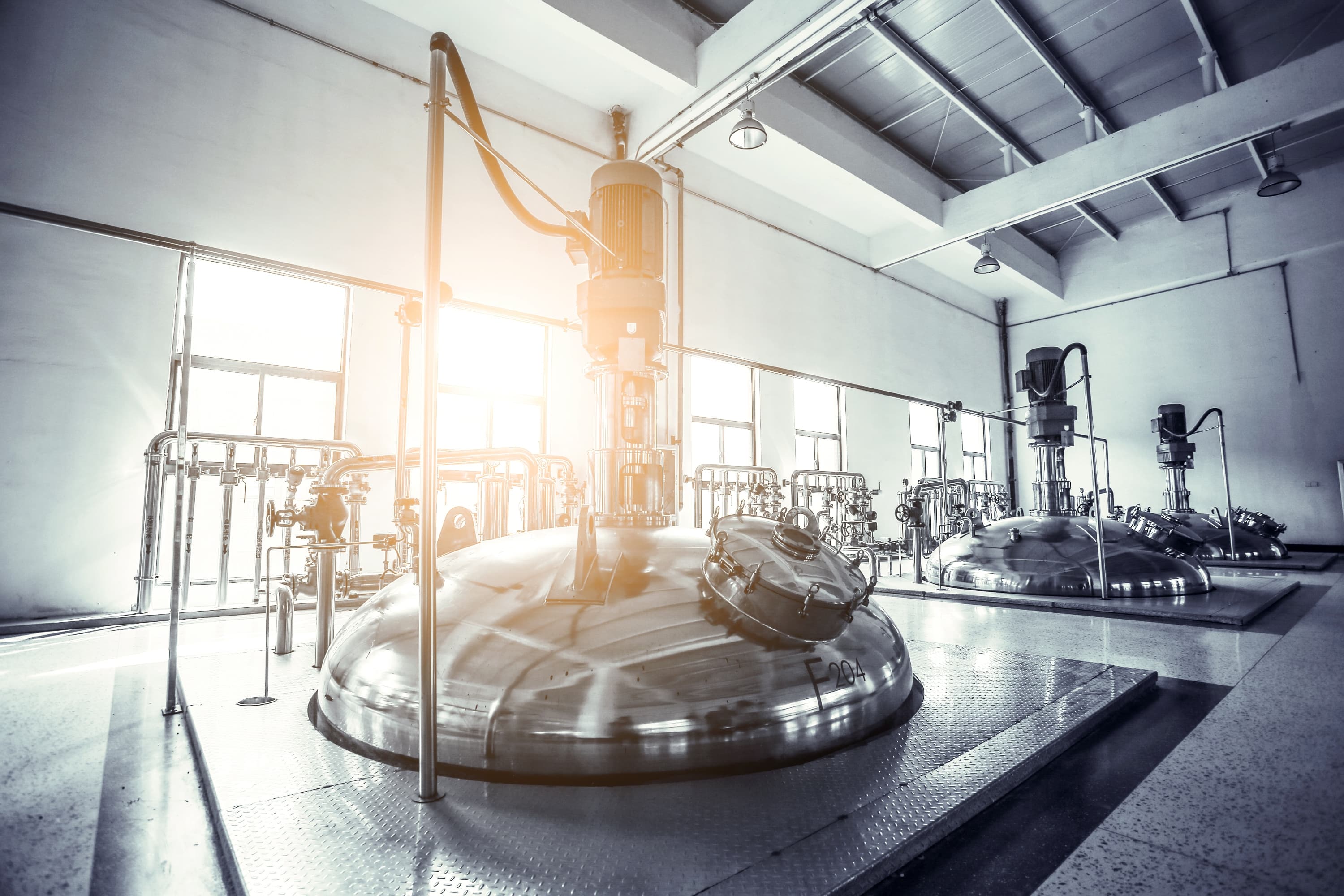 Industrial processing room with large stainless steel reactors, piping, and pumps; sunlight streams through windows, reflecting off metal surfaces.