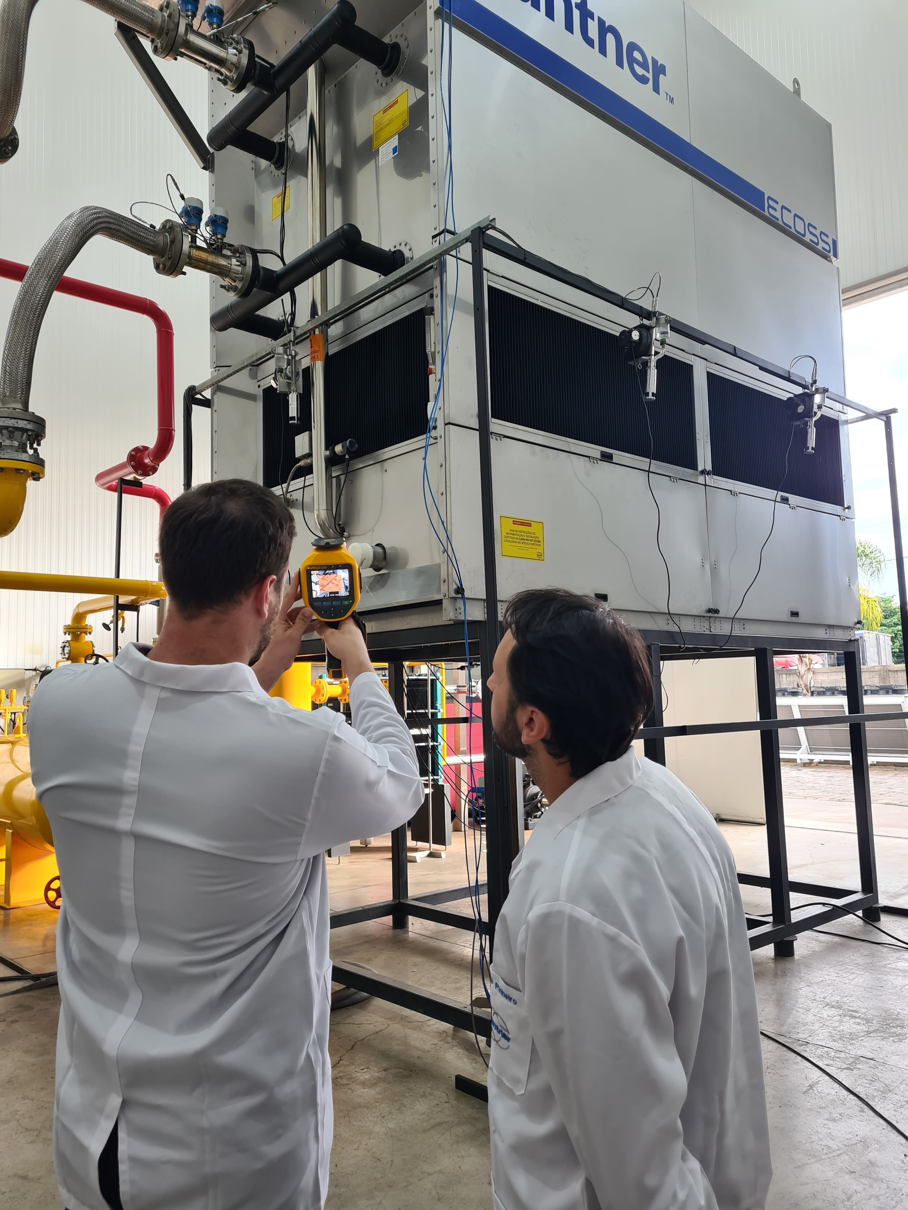 Two technicians in white lab coats inspect a large industrial cooling unit, using a handheld infrared camera.