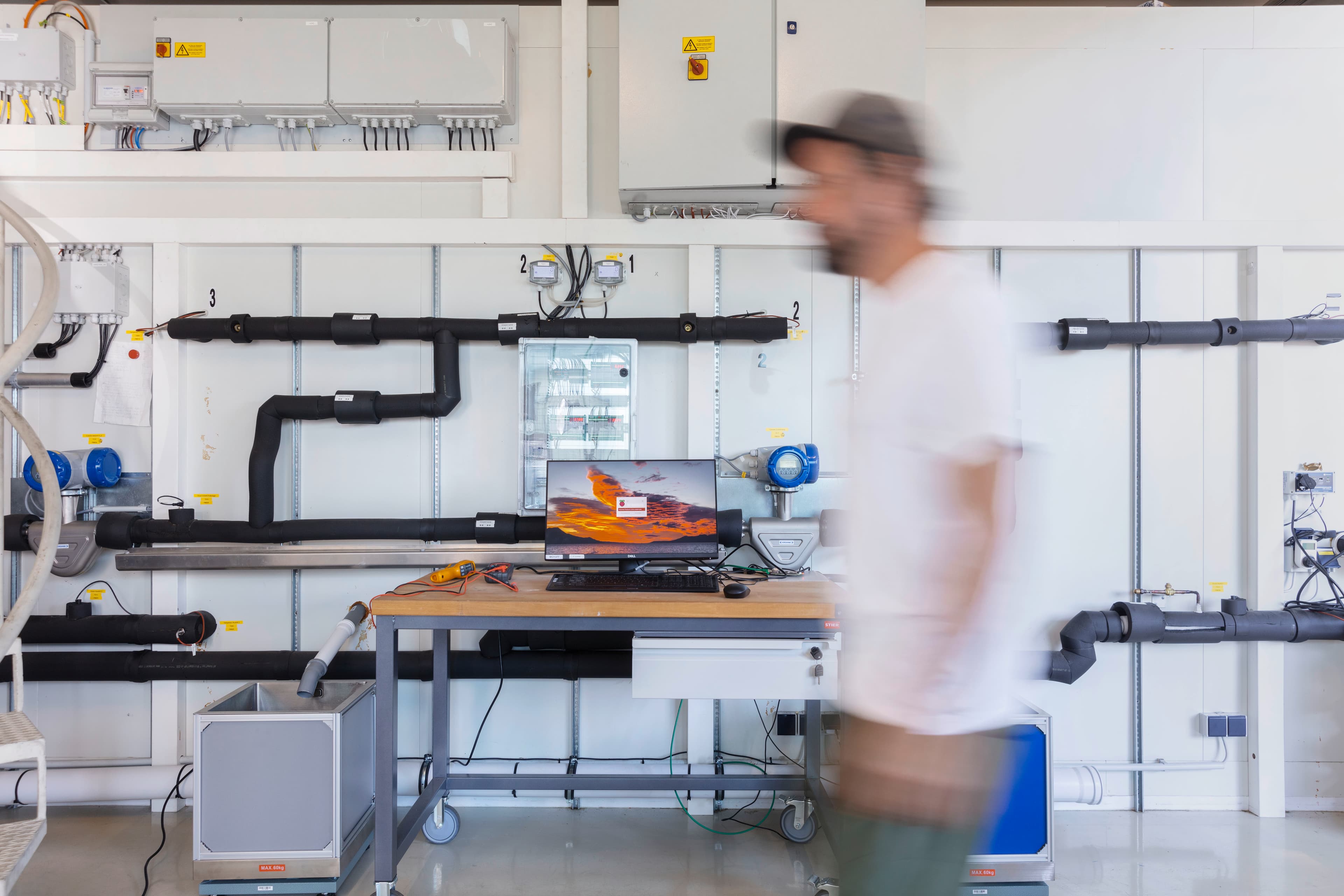 Blurred figure walks past a laboratory workstation with a monitor showing a sunset, surrounded by black insulated pipes and white wall panels.