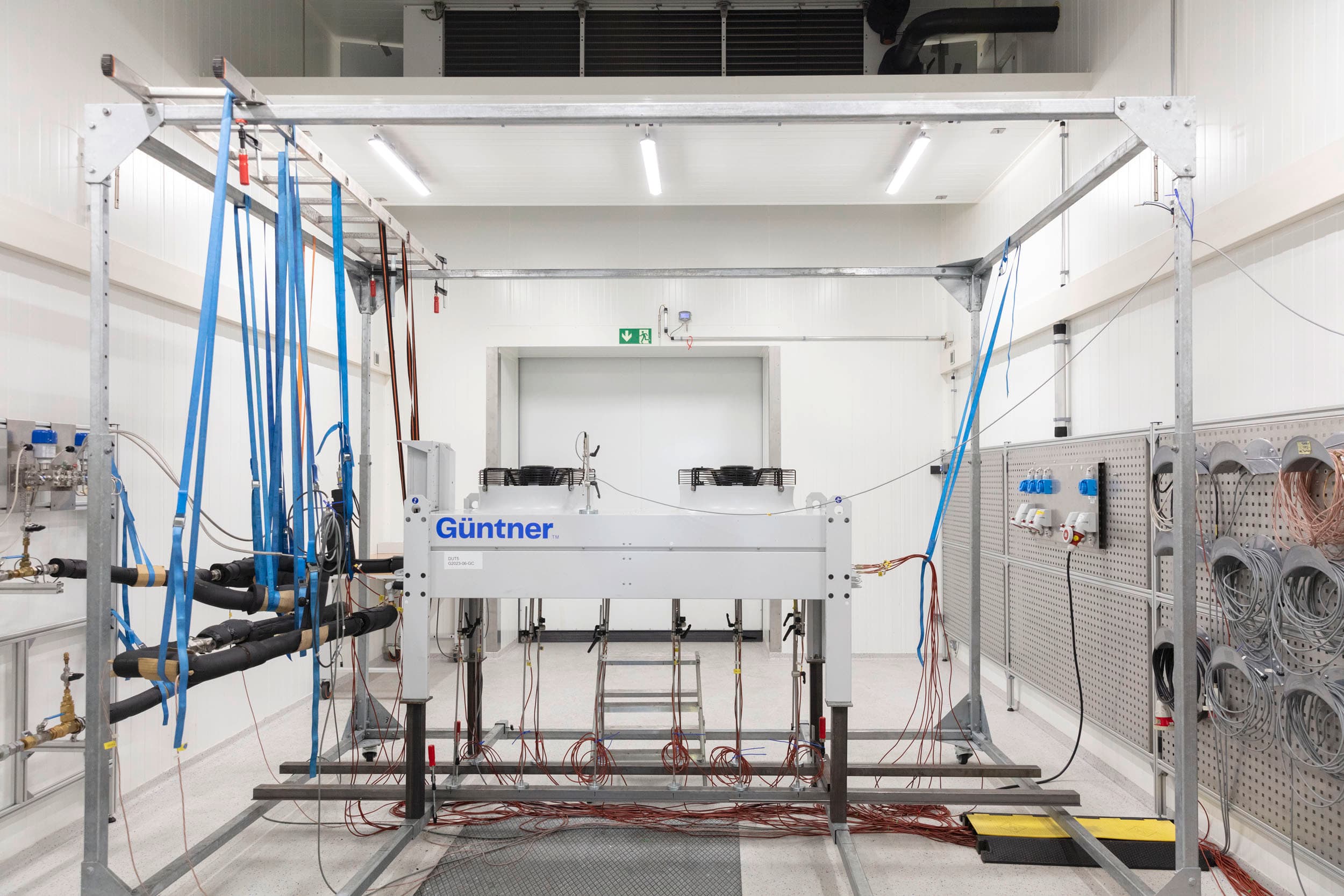 Industrial test rig in a bright white lab; a large white Günttner machine on a metal frame, with blue straps, cables, and a pegboard with coils on the right.