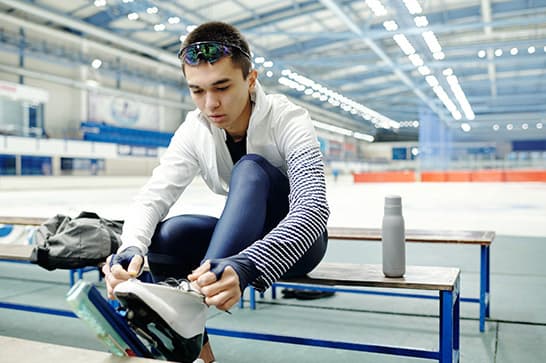 Person sits on a bench at an ice rink, ties skates; wearing a white jacket with striped sleeves and sunglasses on head, with a water bottle nearby.