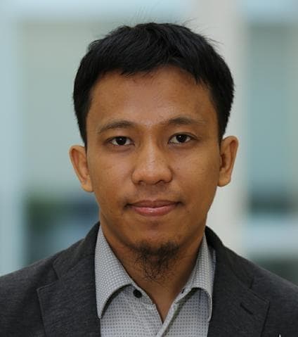 Bust portrait of a person with short black hair and a goatee, wearing a gray blazer and light patterned shirt, neutral expression, blurred background.