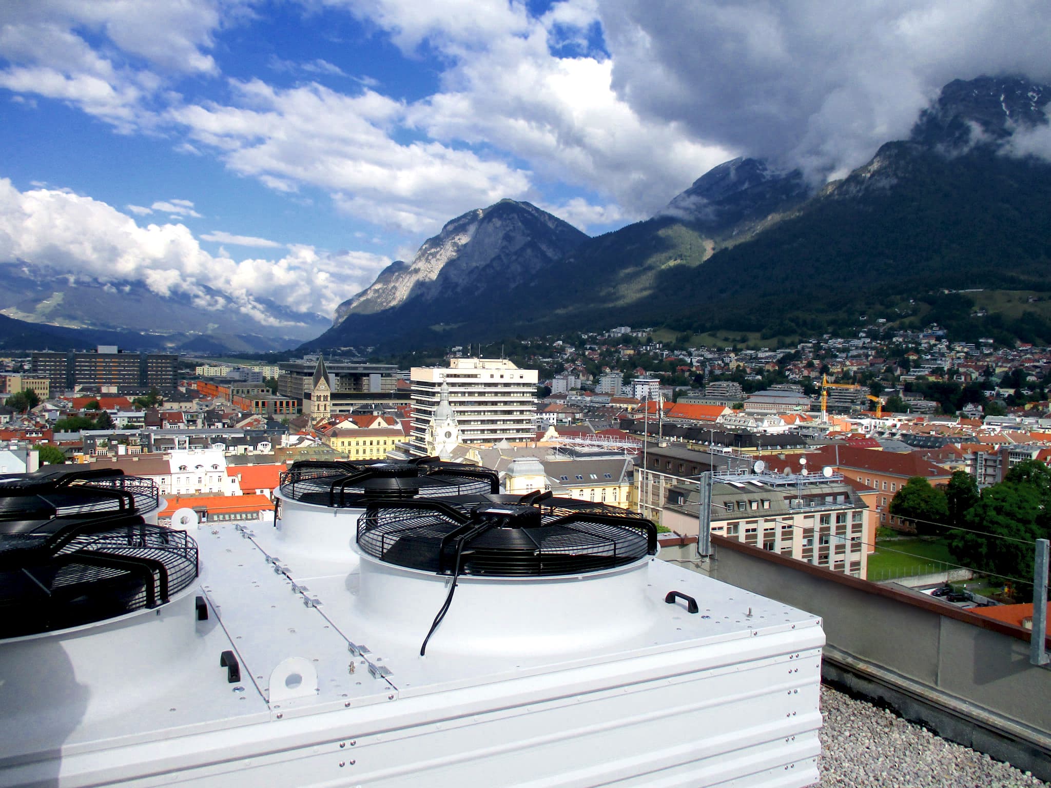 Rooftop HVAC units and fans in the foreground; a European city with red-tiled roofs and a church spire, set against green mountains and a blue sky.