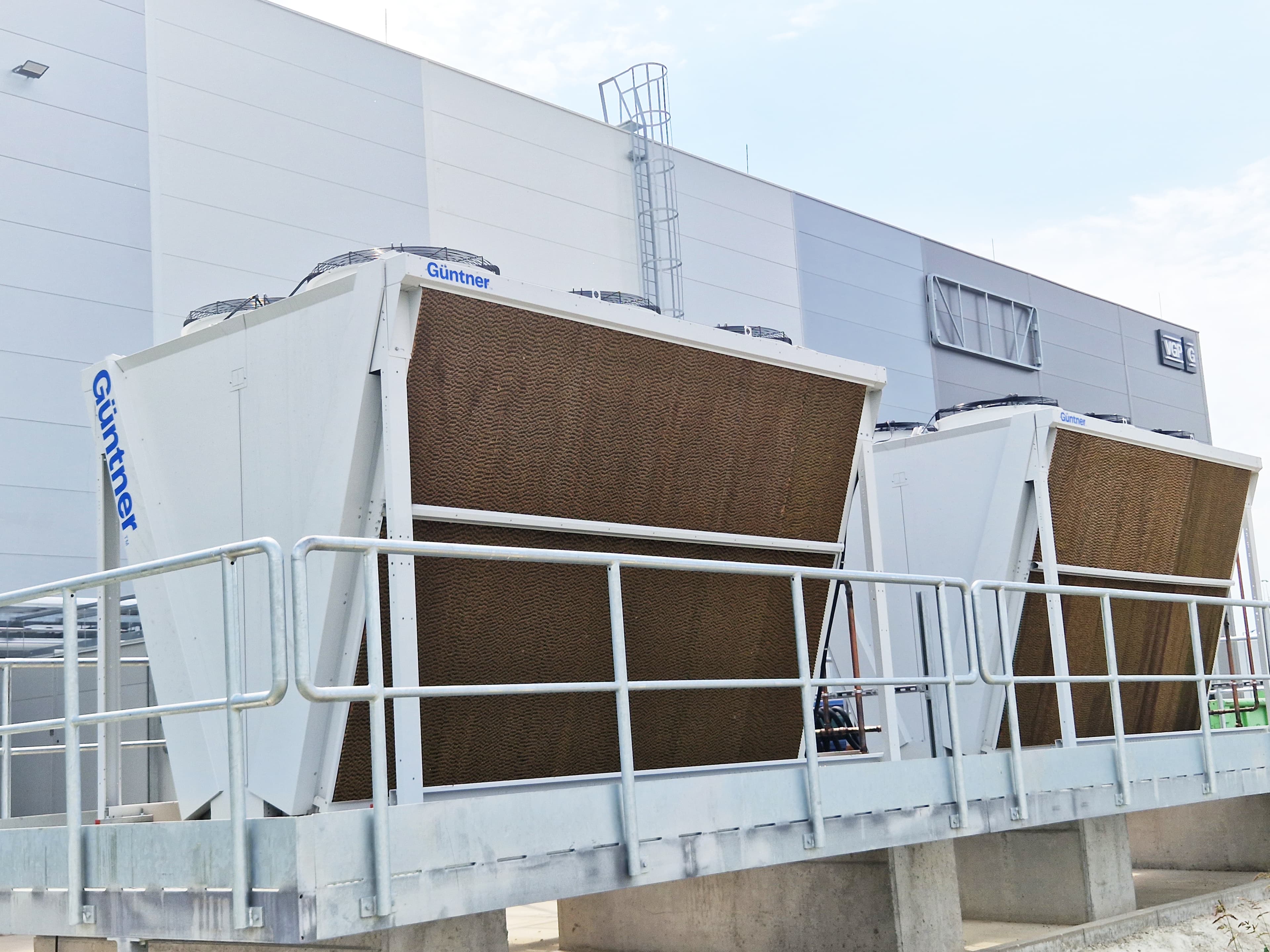 Two large white cooling units with brown finned heat exchangers labeled 'Guntner' on a raised platform with railing beside a gray industrial building.