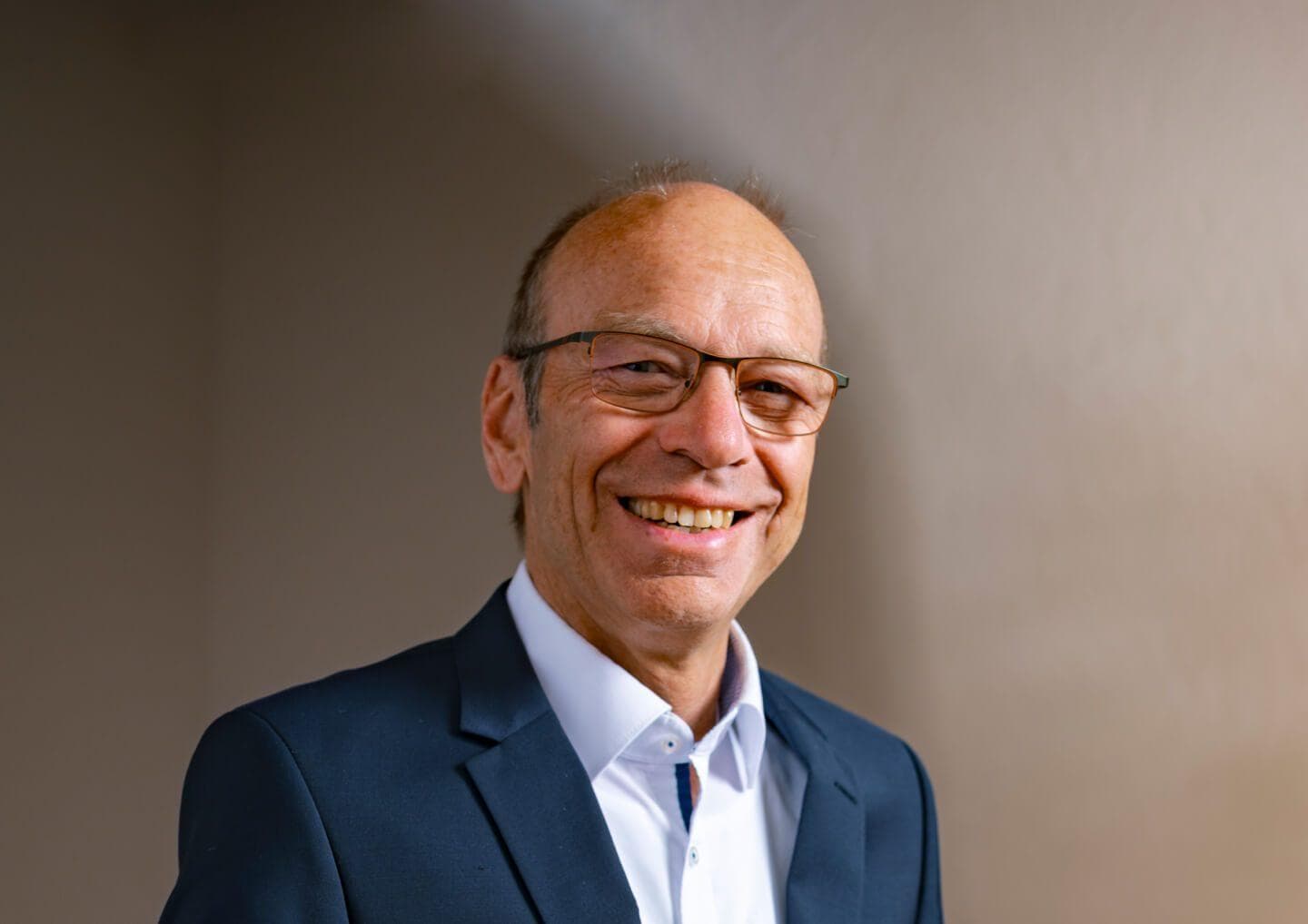 Eine Person mit Brille, dunklem Anzug und weißem Hemd, die freundlich lächelt, vor einem hellen, neutralen Hintergrund.