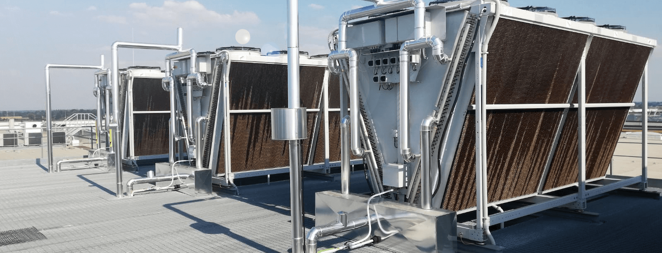 Rooftop cooling system with large brown finned heat exchangers and silver piping on a metal deck under a blue sky.