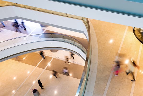 A modern shopping mall atrium viewed from above, featuring sweeping curved white walkways, glass railings, and light tiled floors with blurred shoppers.