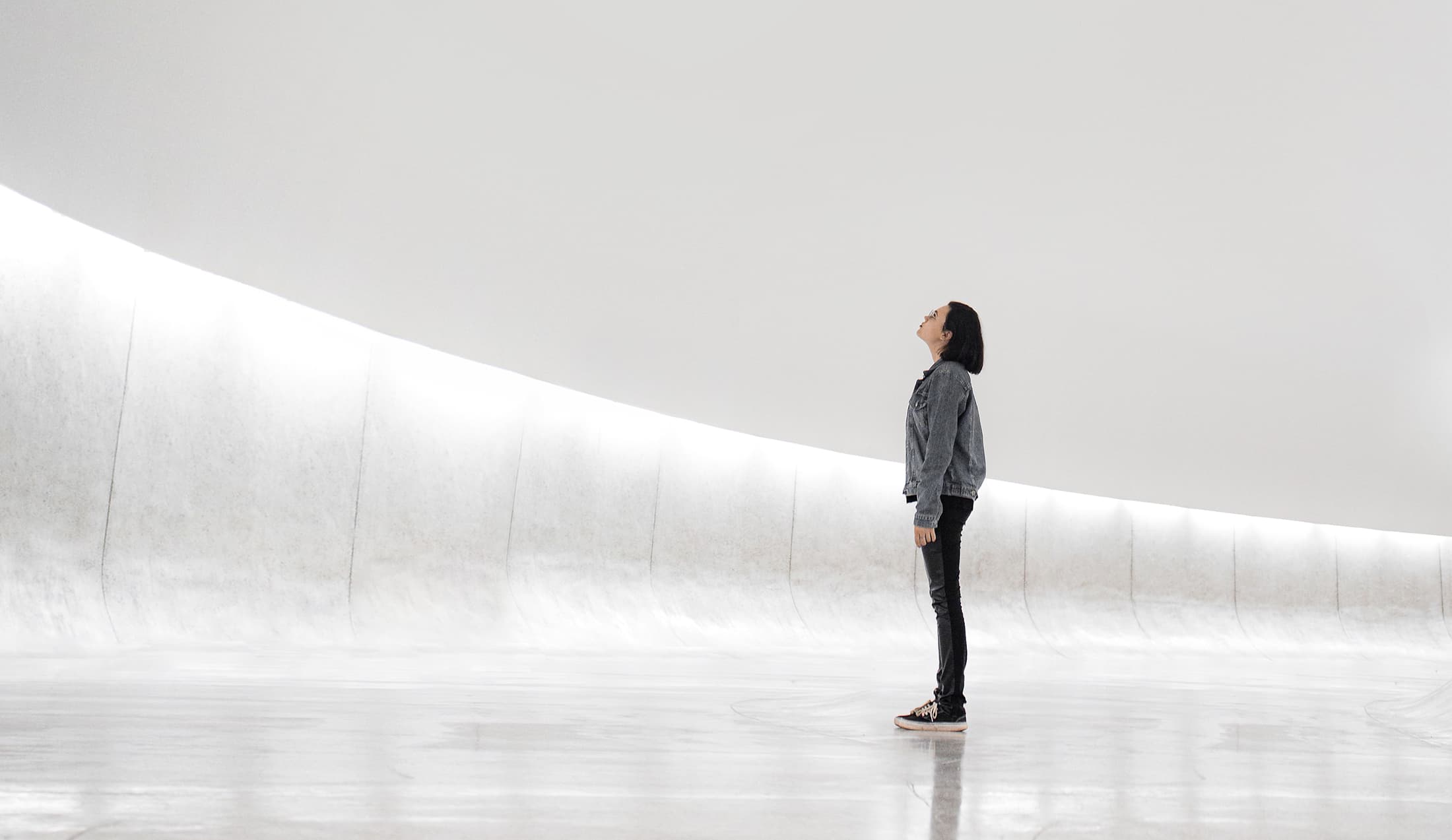 A person in a denim jacket and black pants stands in a large, curved white gallery space, looking up at a bright, minimalist ceiling.